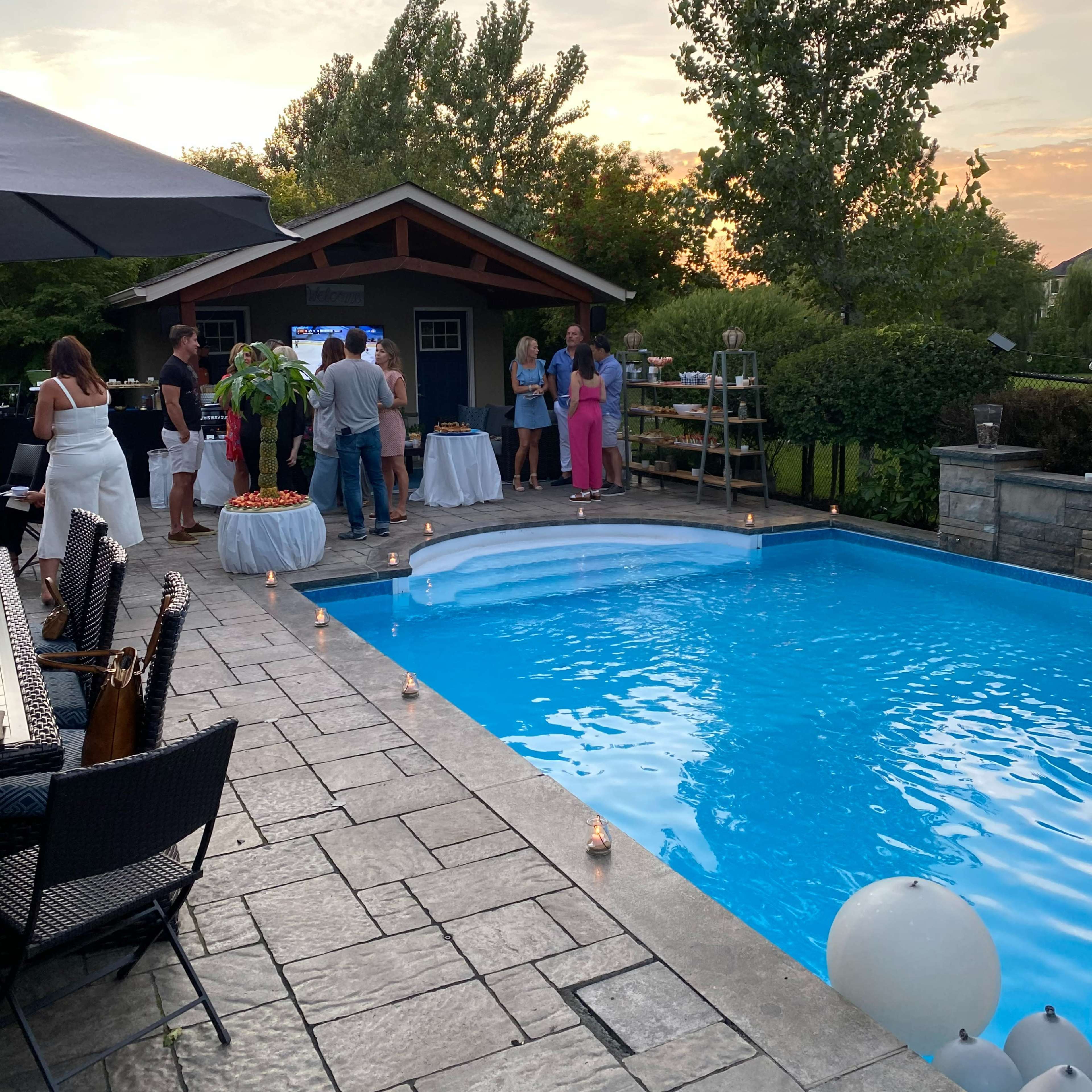 A group of people gather around a poolside area with tables and candles as the sun sets in the background.