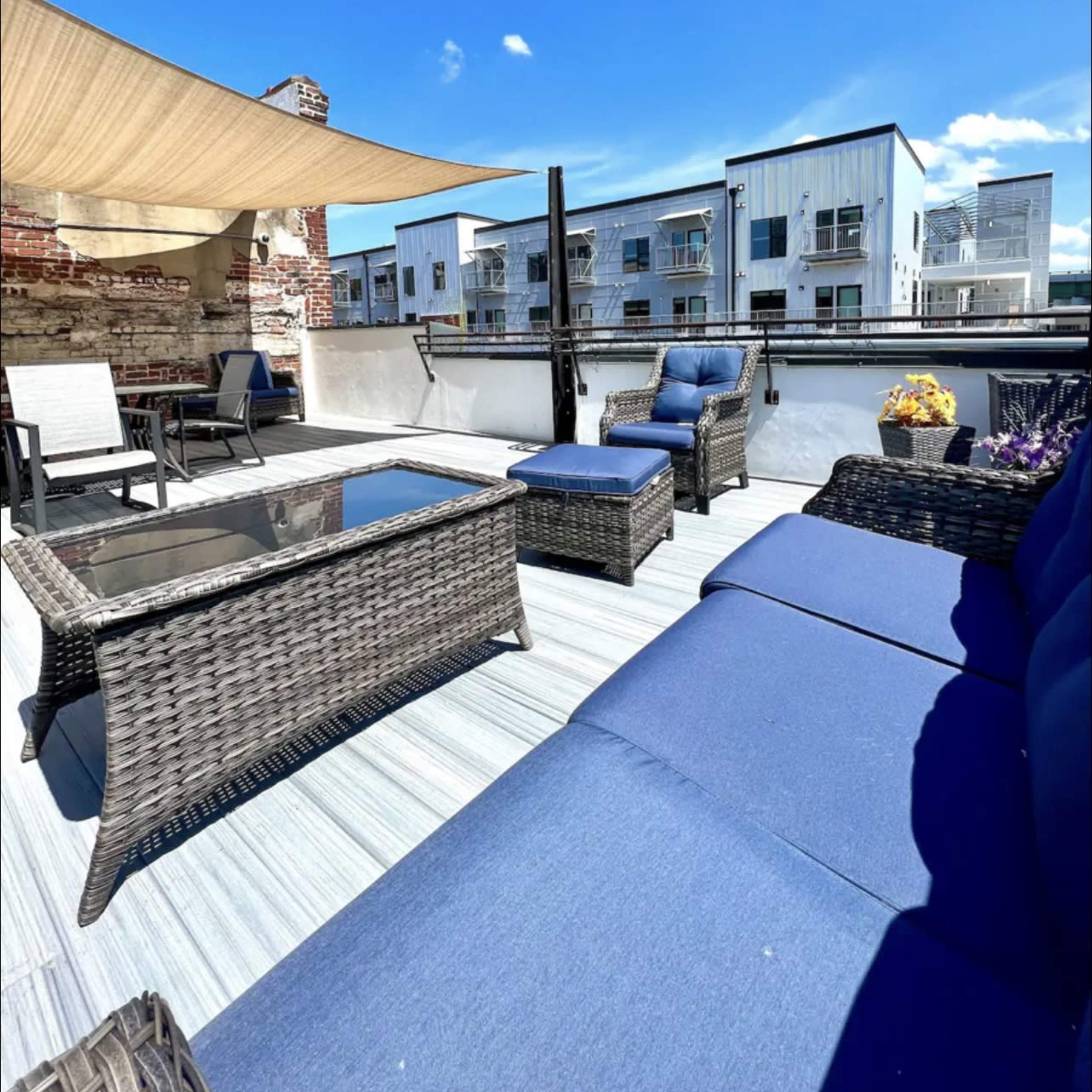 A rooftop patio with wicker furniture, a shaded area, and a view of nearby modern buildings under a clear blue sky.