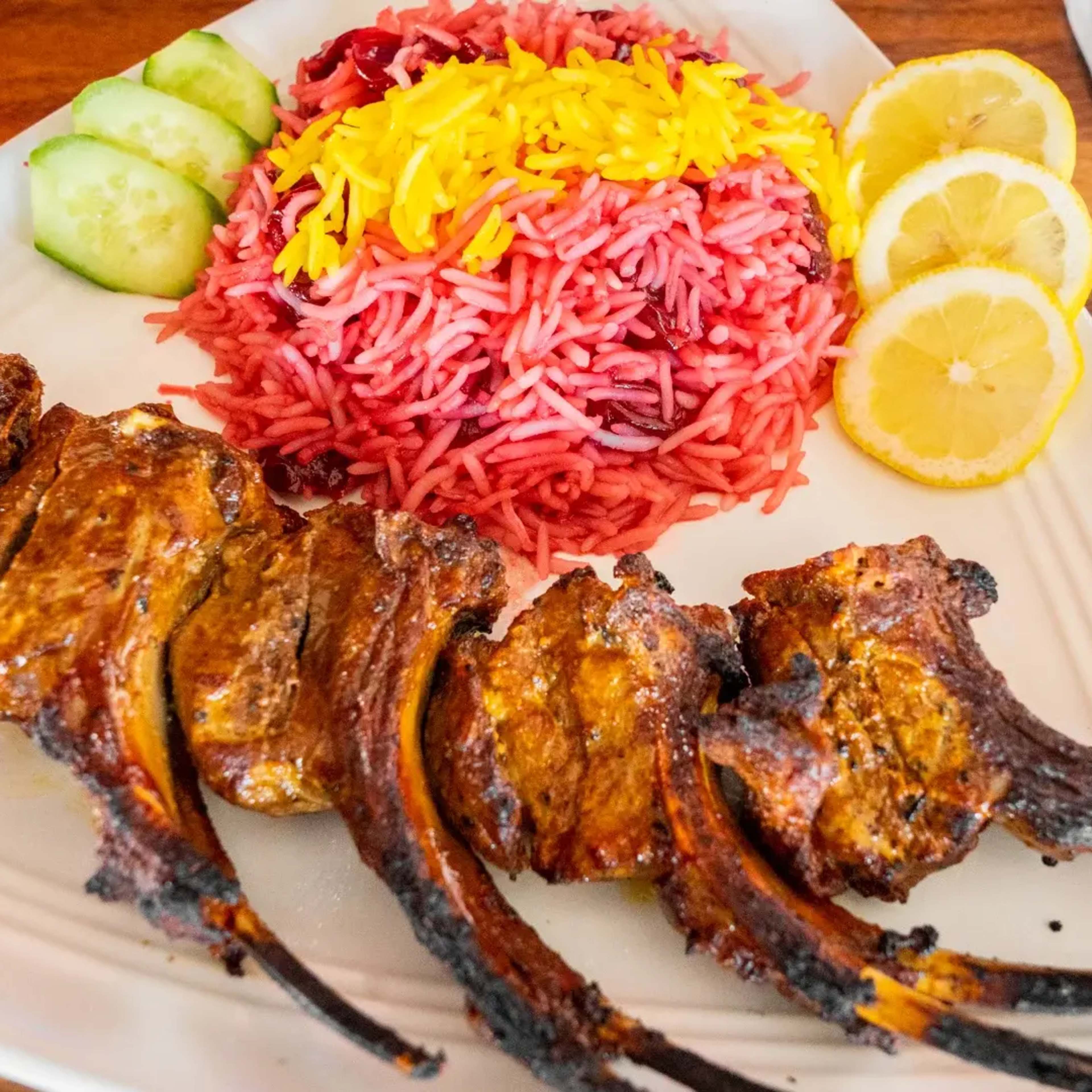 A plate of grilled lamb chops alongside a serving of colorful rice topped with yellow strands and garnished with cucumber slices and lemon wedges.