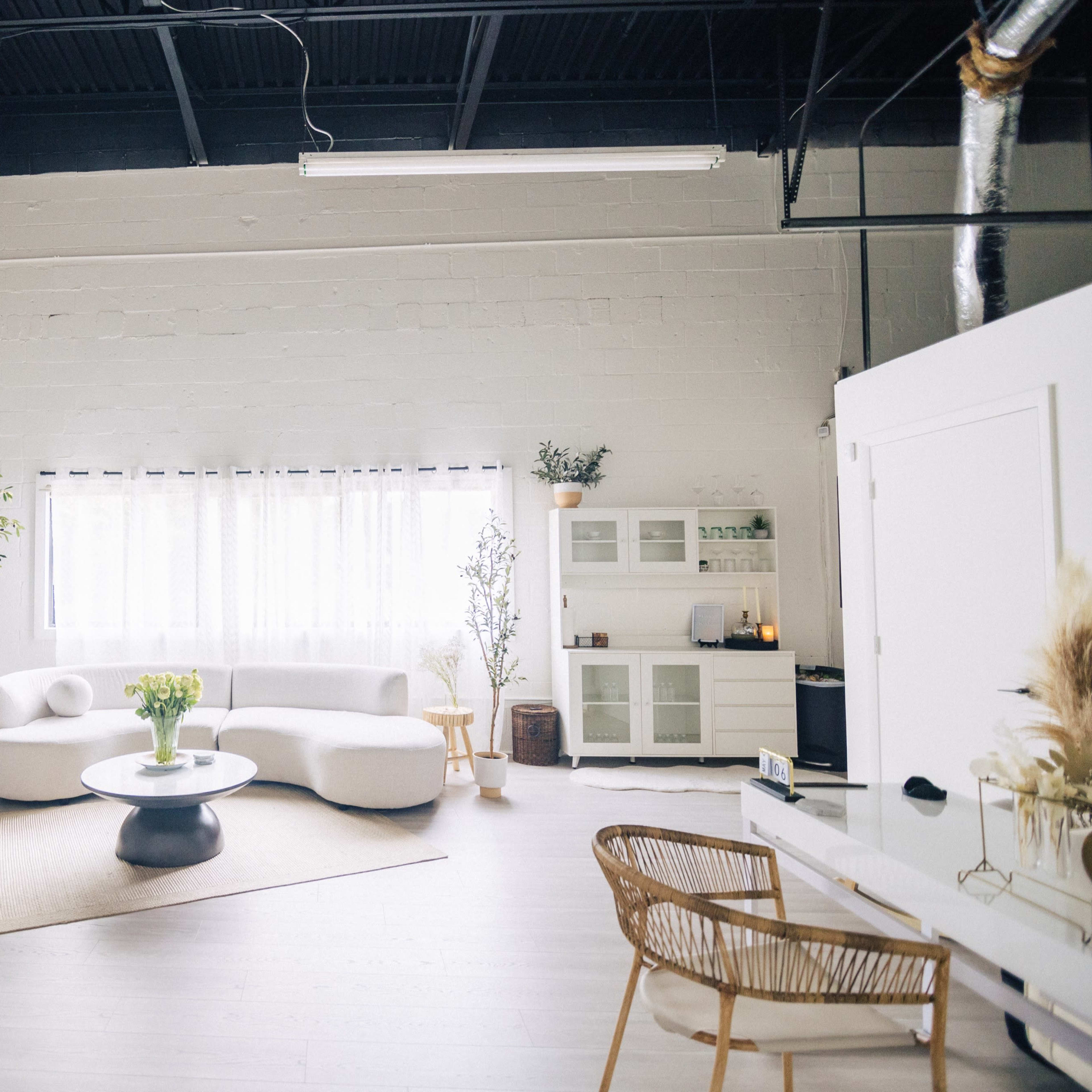 A spacious, well-lit studio with a white sofa, round coffee table, a large plant, and minimalist decor, including a shelf with various items.