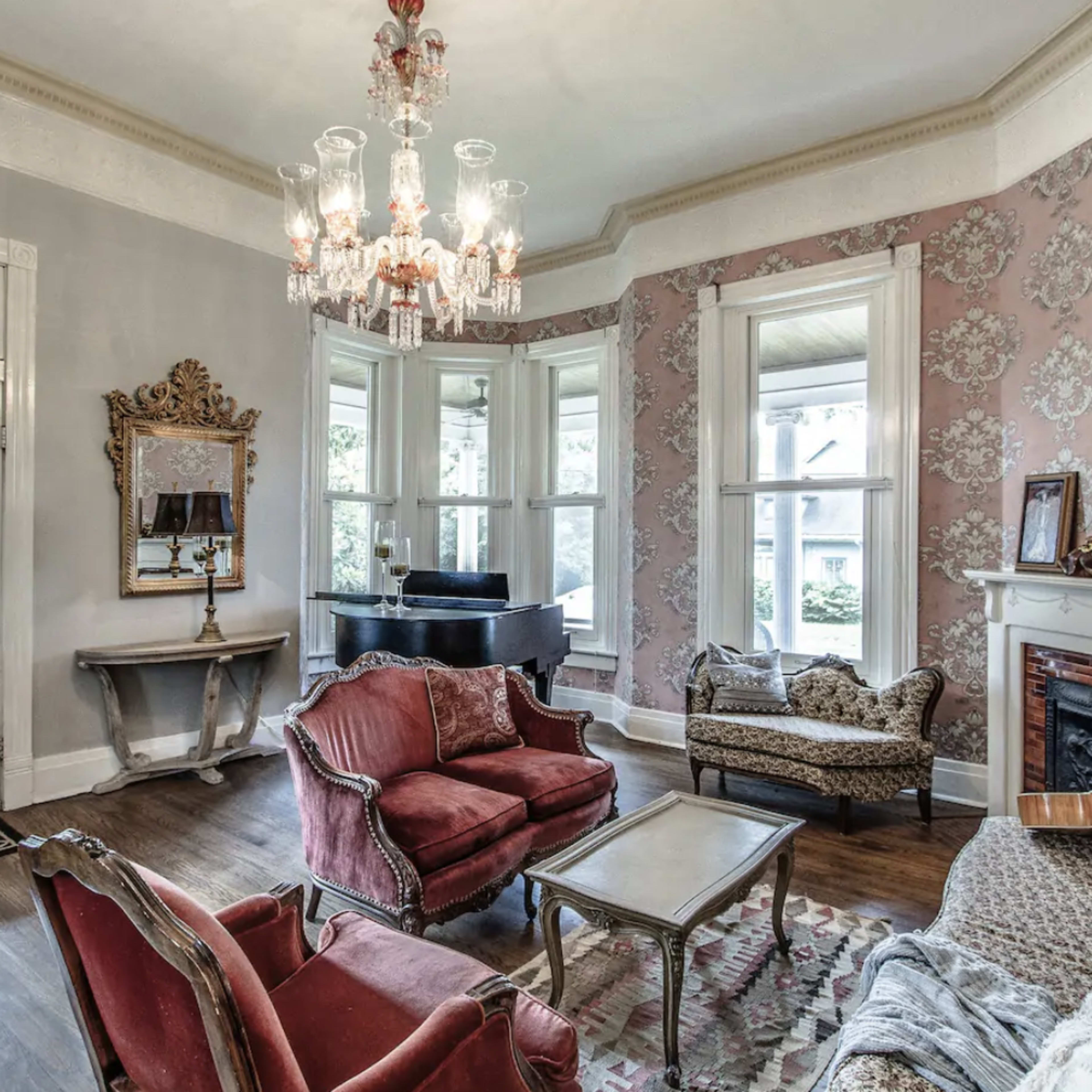 The living room features vintage furnishings, a chandelier, and large windows that provide natural light.