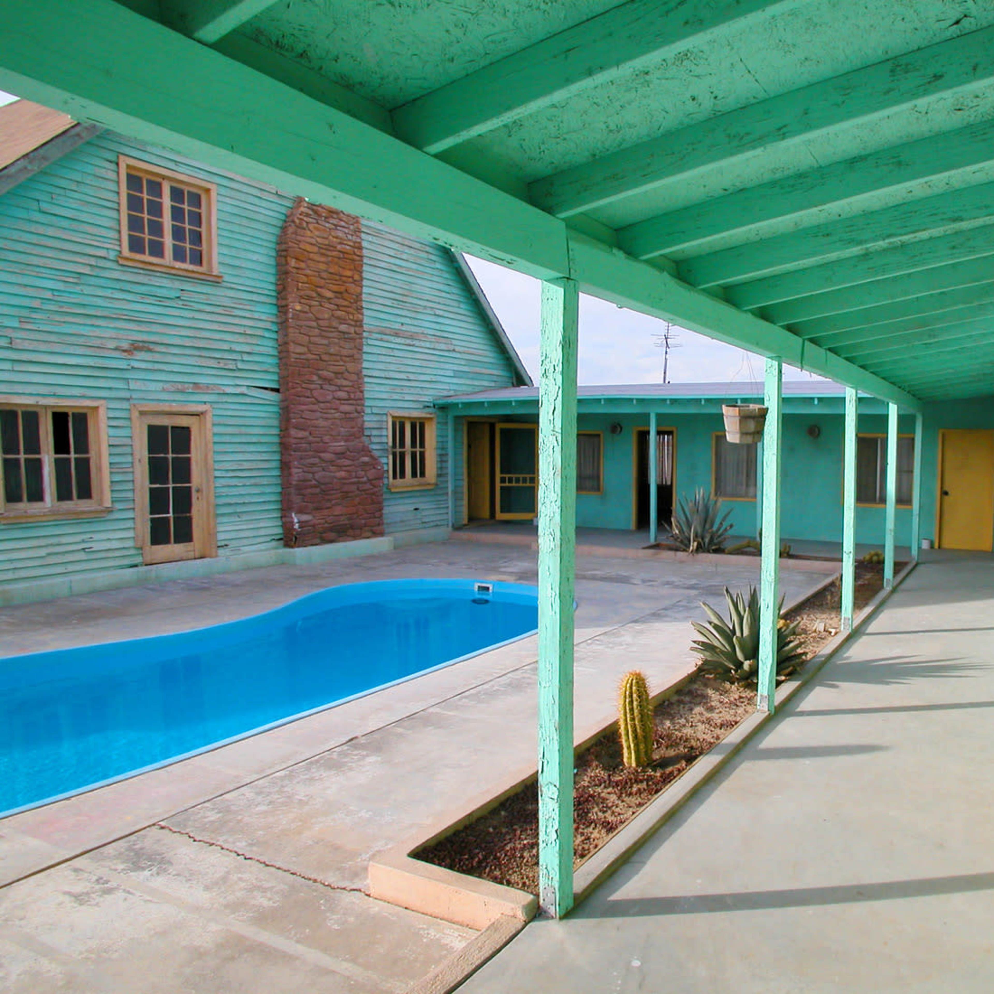A turquoise-colored corridor runs alongside a swimming pool surrounded by a courtyard and buildings with a vintage design.