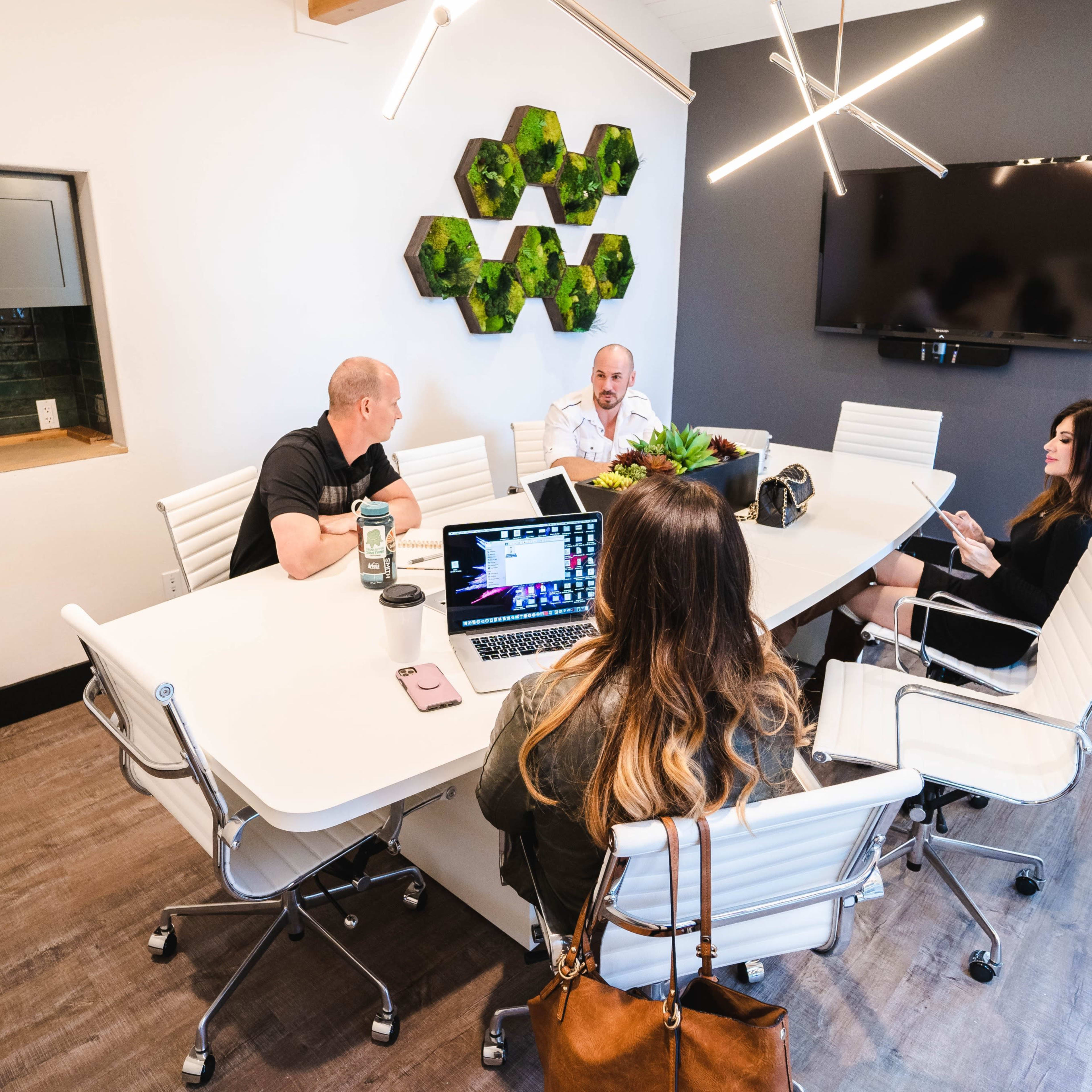 A group of five people are seated around a modern conference table in a well-lit meeting room, with a large screen and decorative wall elements in the background.