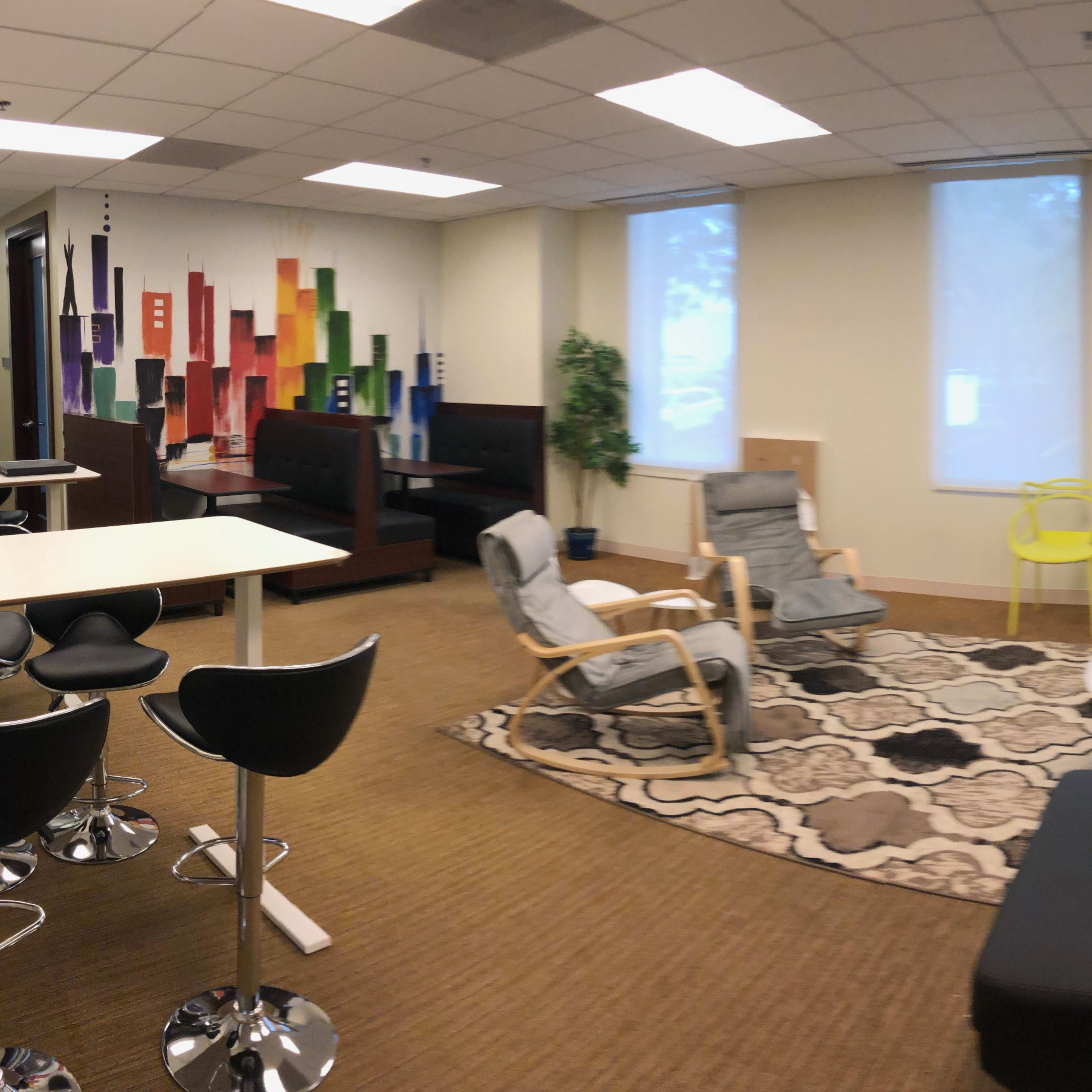 A spacious lounge area featuring a mix of seating arrangements, including black sofas, colorful chairs, and a round table on a patterned rug.