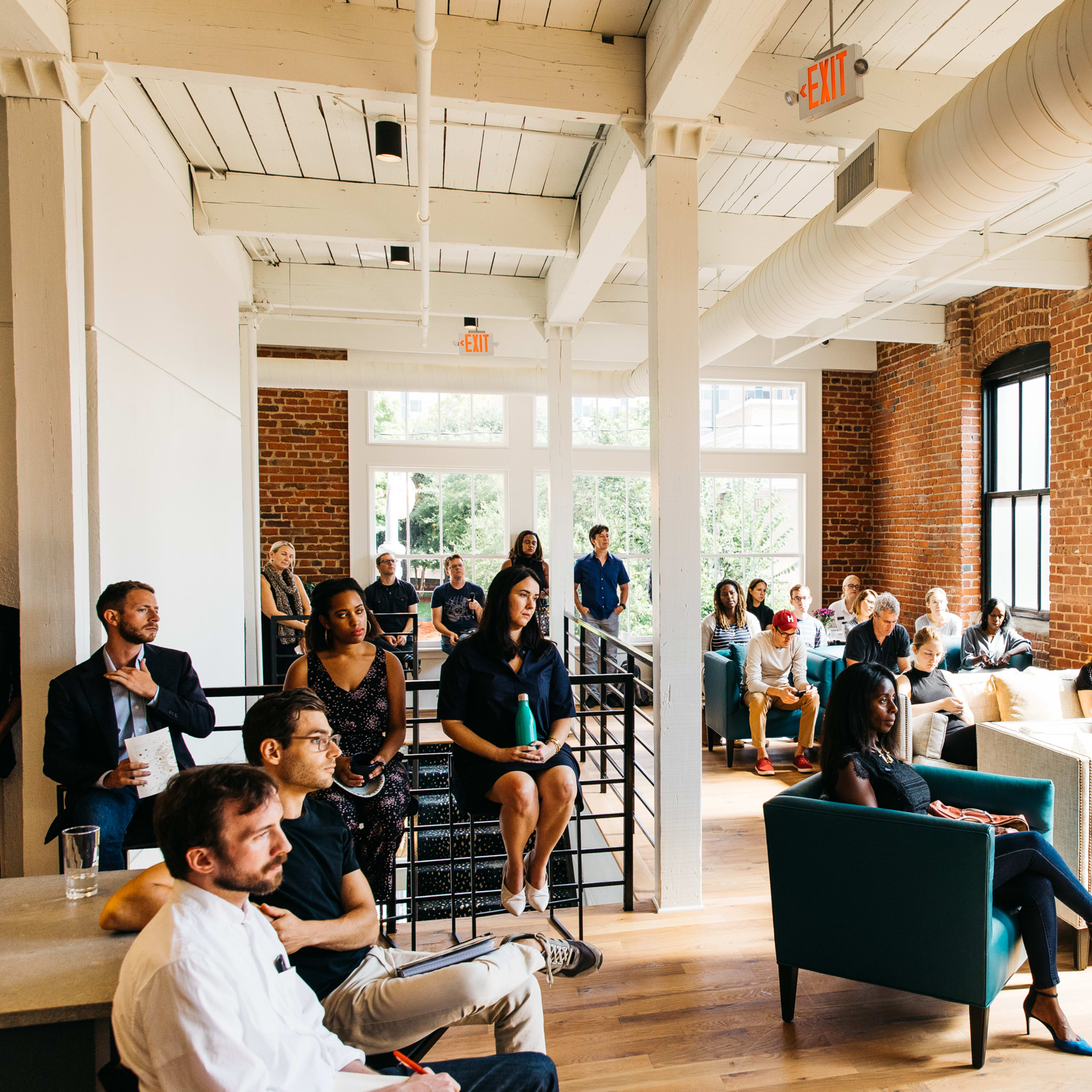 A diverse group of people is seated and standing in a bright, open brick-walled room with large windows, engaged in a presentation or discussion.