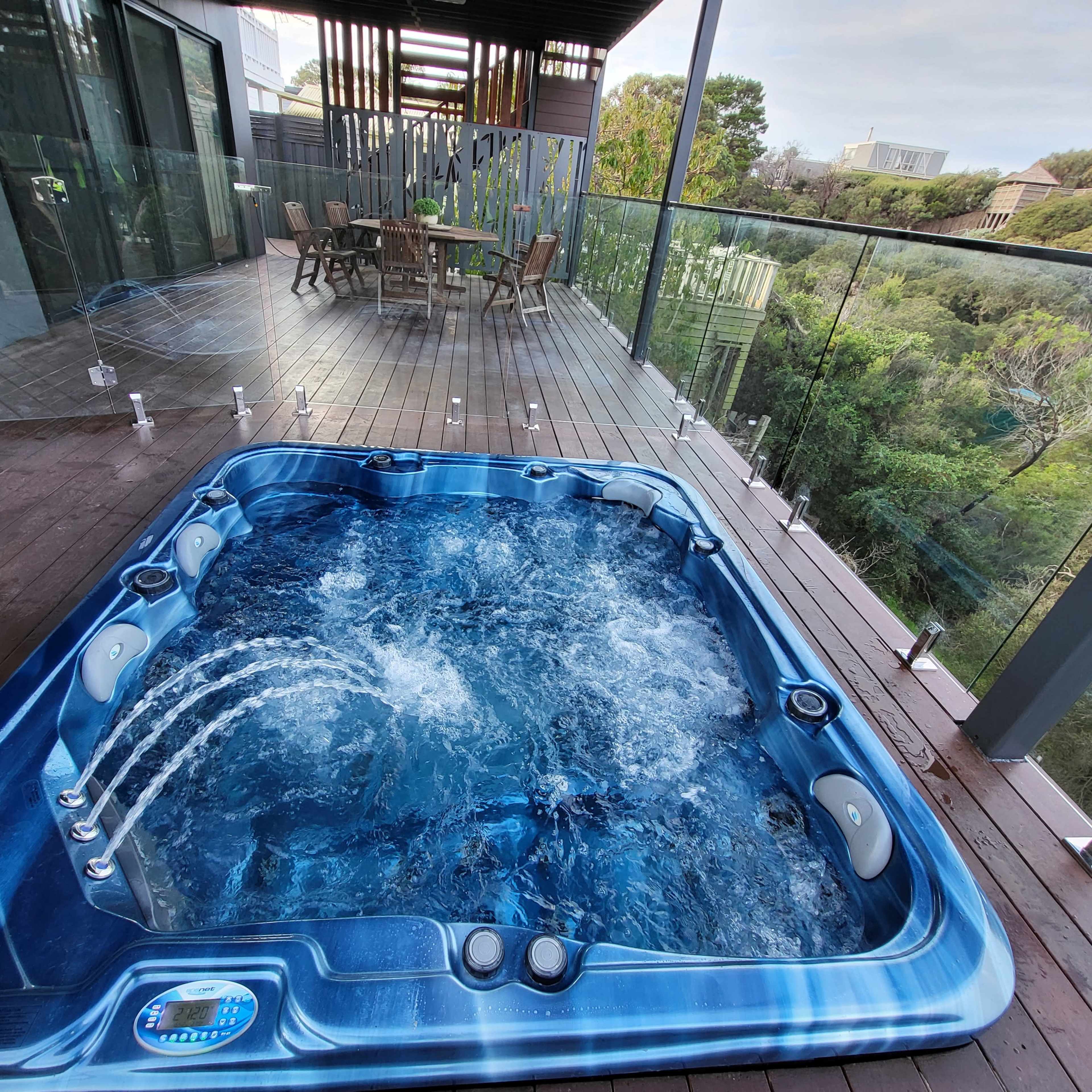A hot tub with jets is located on a wooden deck alongside large glass windows overlooking greenery.