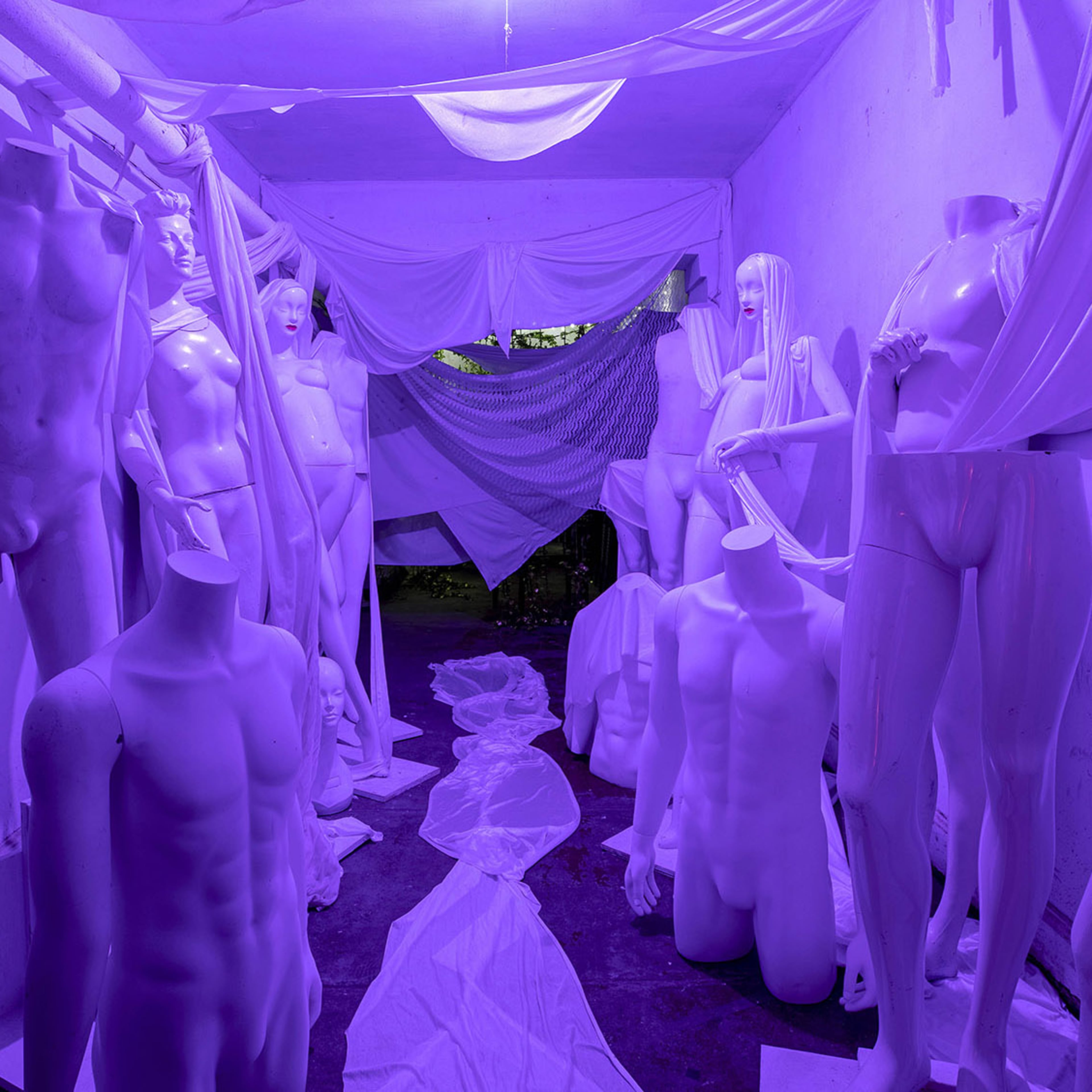 A narrow hallway filled with white mannequins draped in sheer fabric under purple lighting.