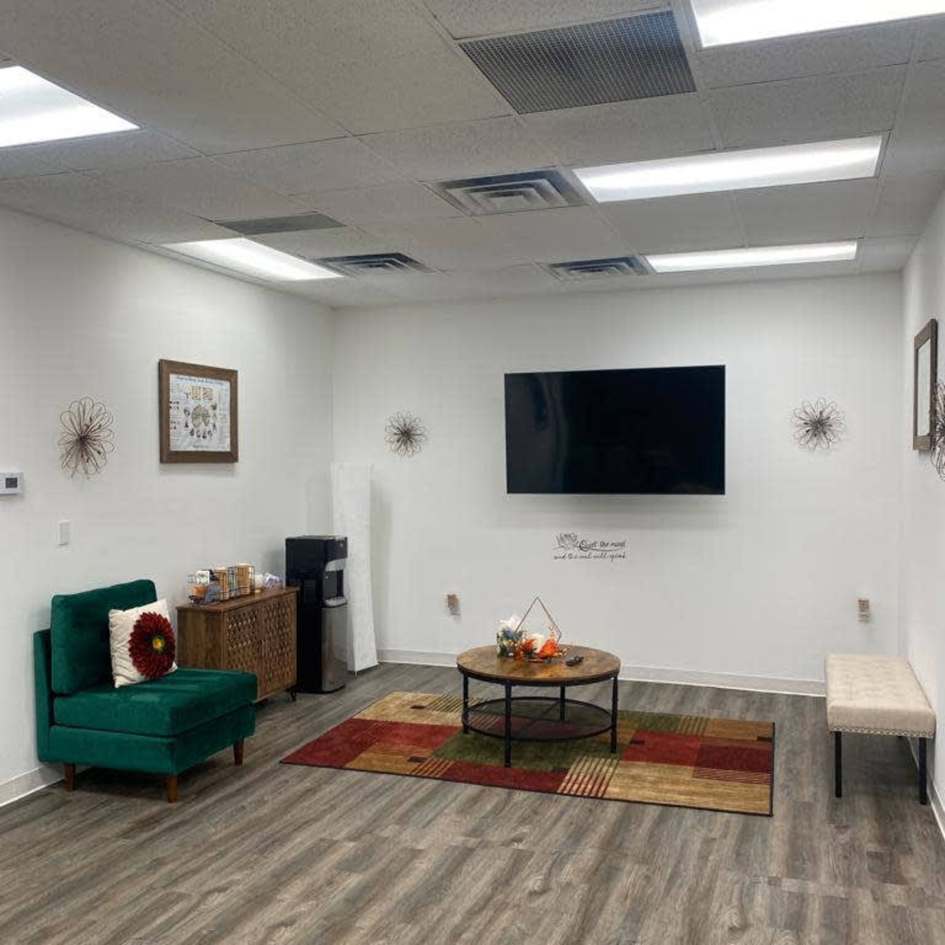 A modern, minimally decorated room featuring a green chair, a wall-mounted television, a round coffee table with a centerpiece, and a patterned area rug on a wood floor.