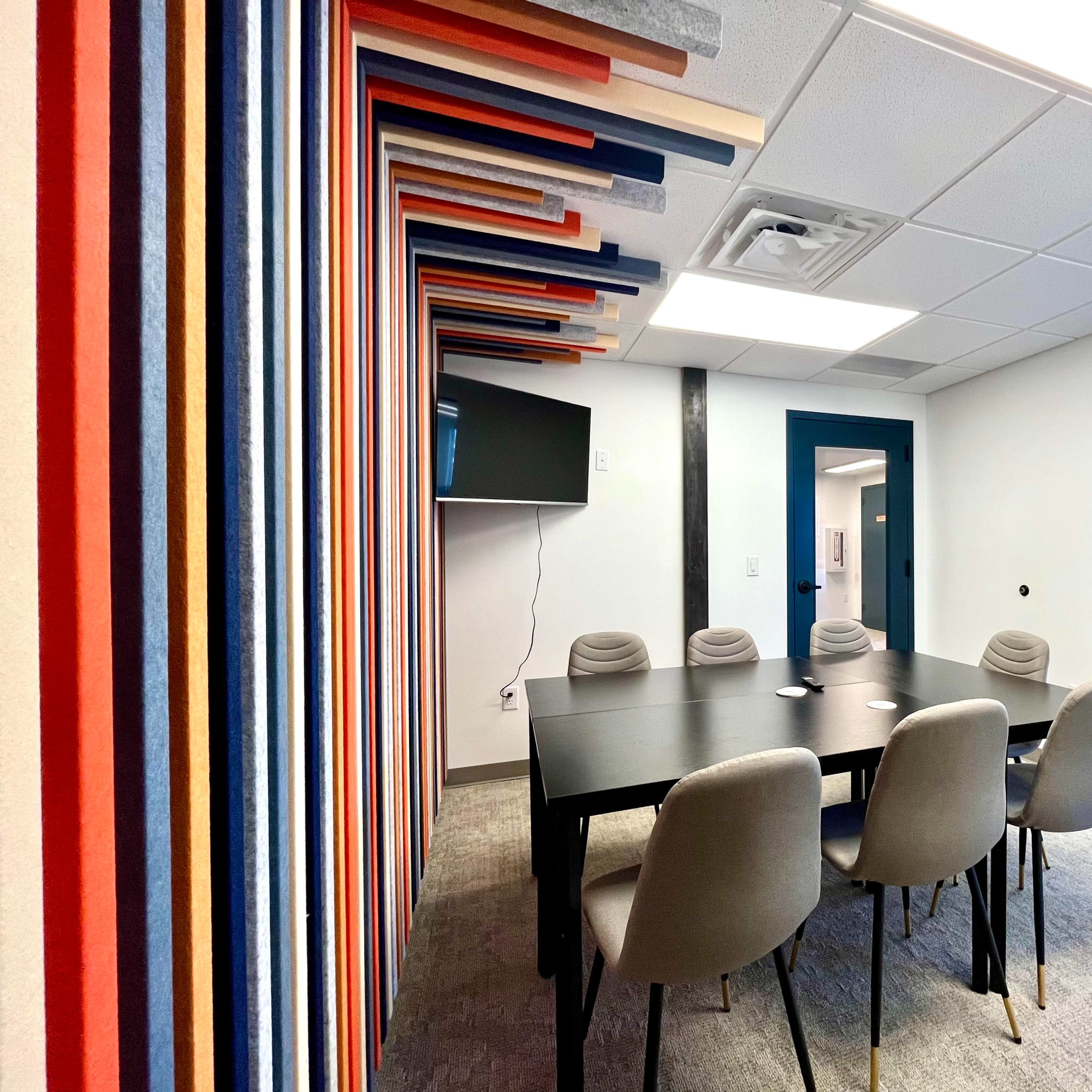 A modern conference room featuring a black table surrounded by gray chairs, with a colorful striped wall and a wall-mounted television.