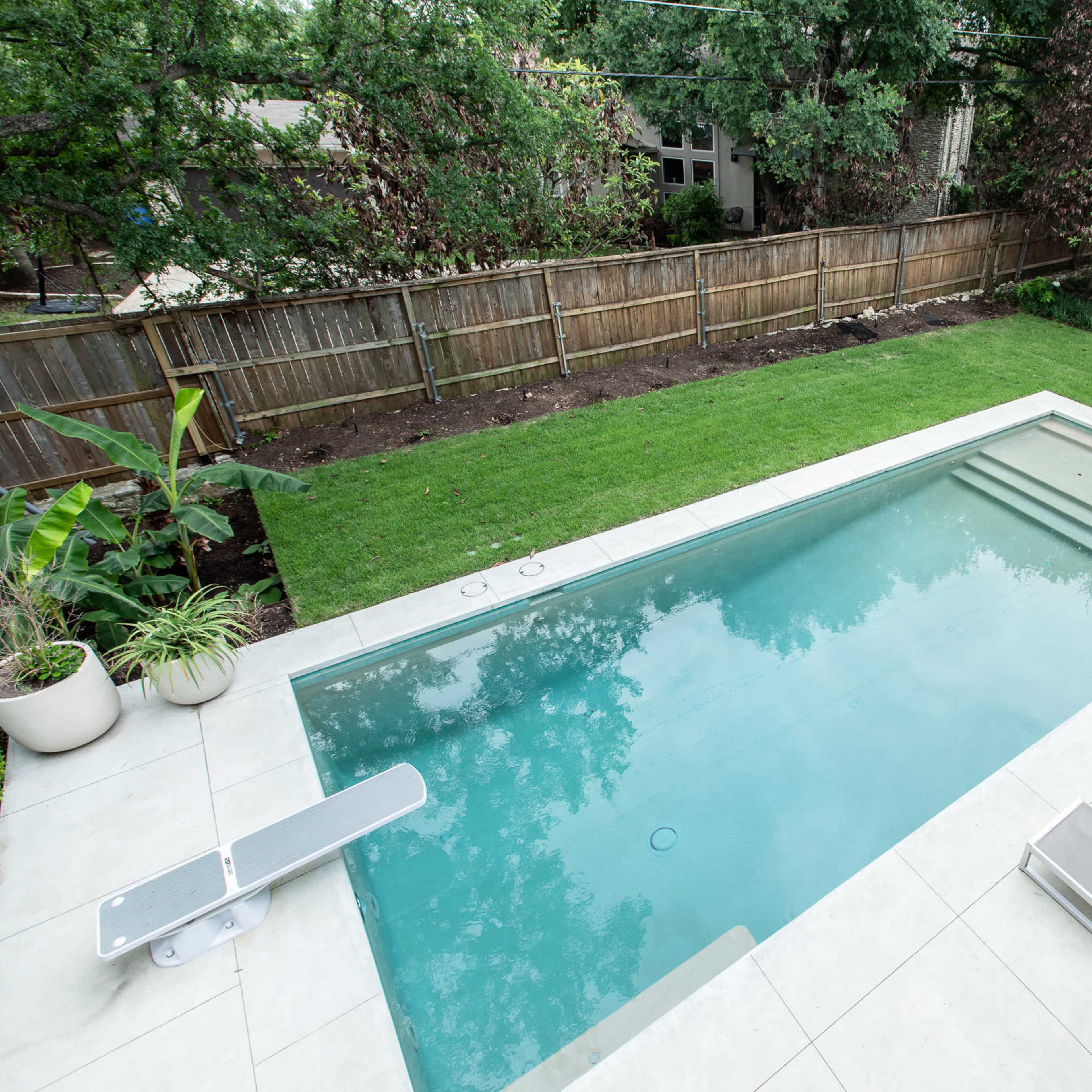 A rectangular swimming pool surrounded by a concrete deck, lounge chairs, and a neatly maintained lawn in a backyard setting.