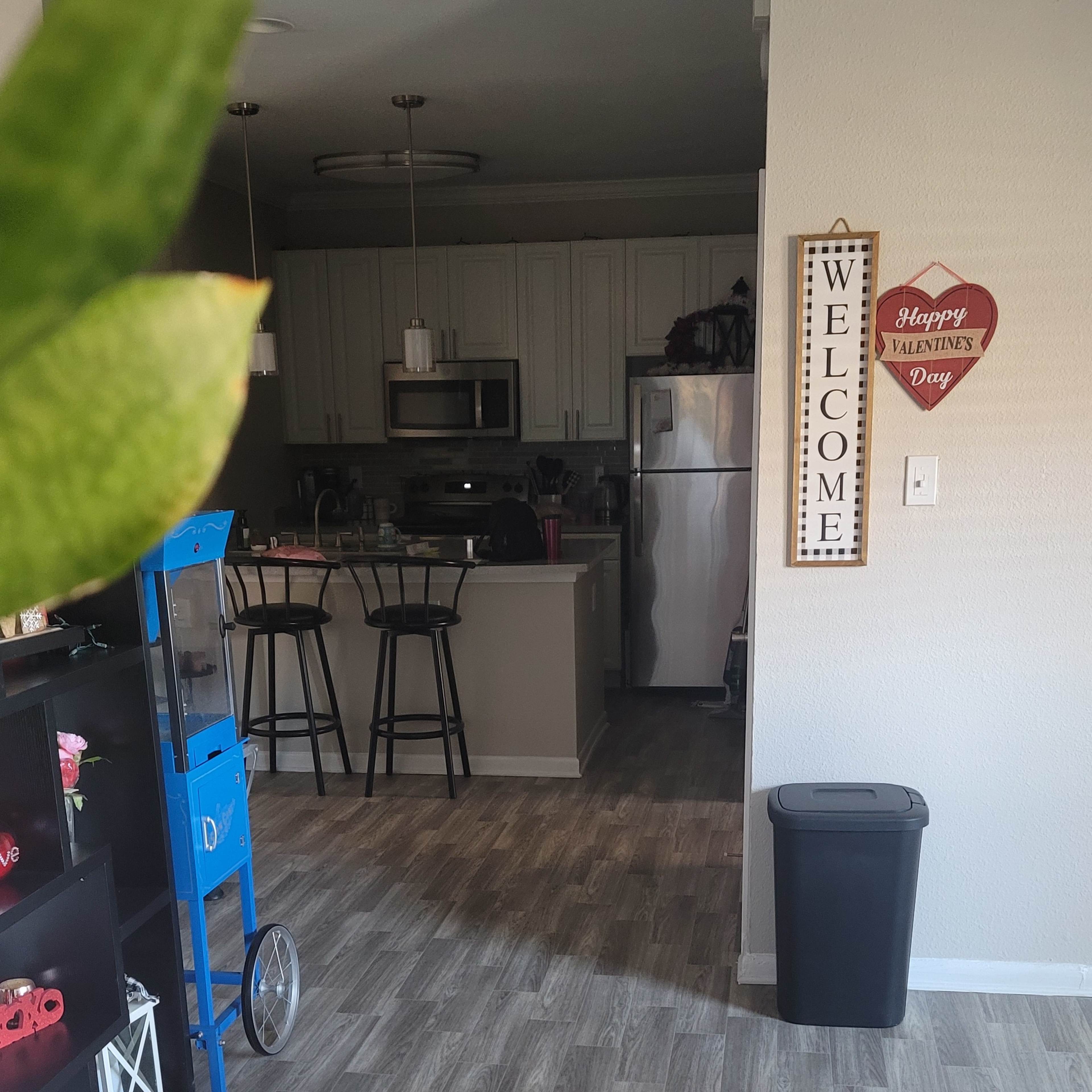 A view of a living area with a welcome sign, a trash can, and a kitchen visible in the background.
