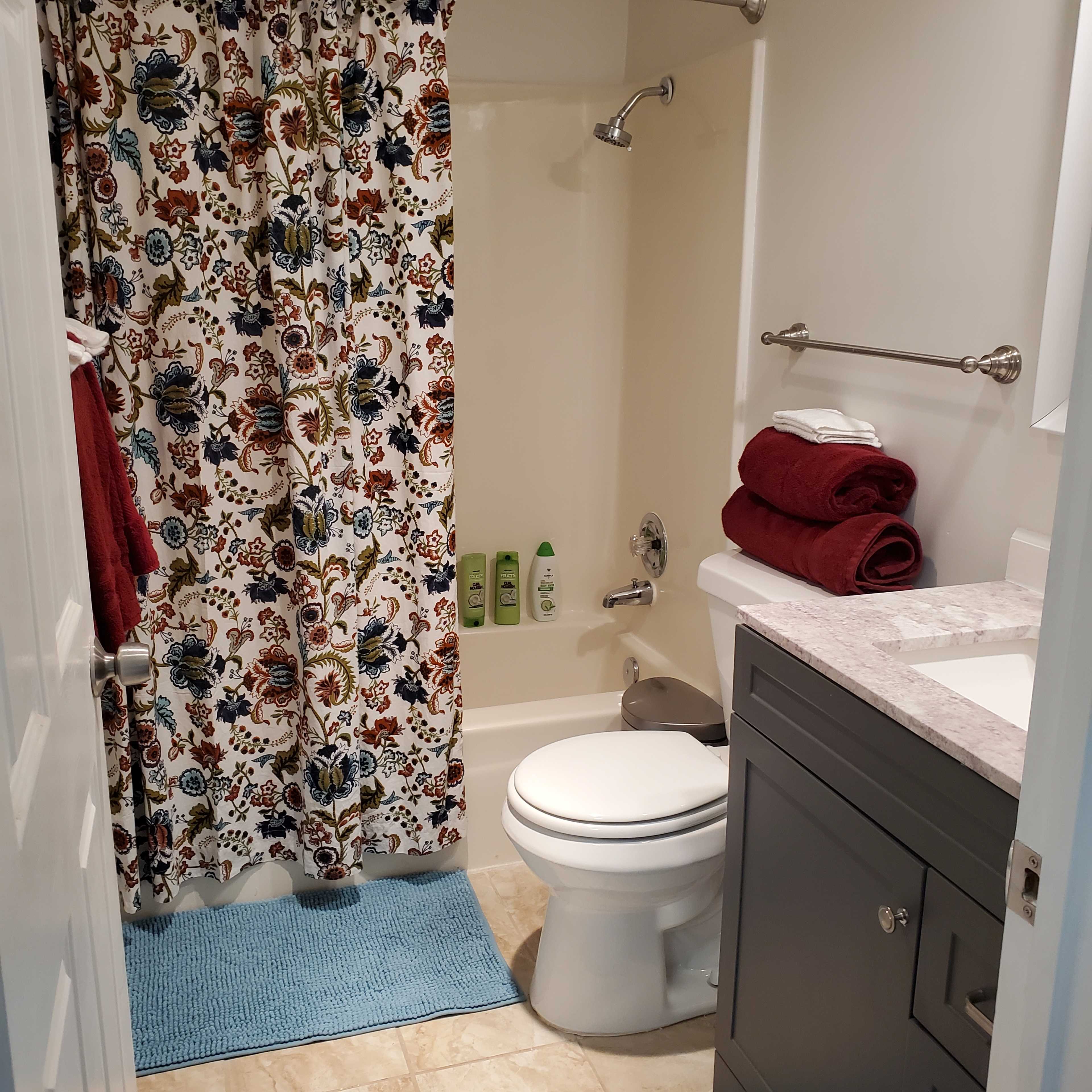 A bathroom with a floral shower curtain, a toilet, a sink with a countertop, towels stacked on a shelf, and a bath mat on the floor.