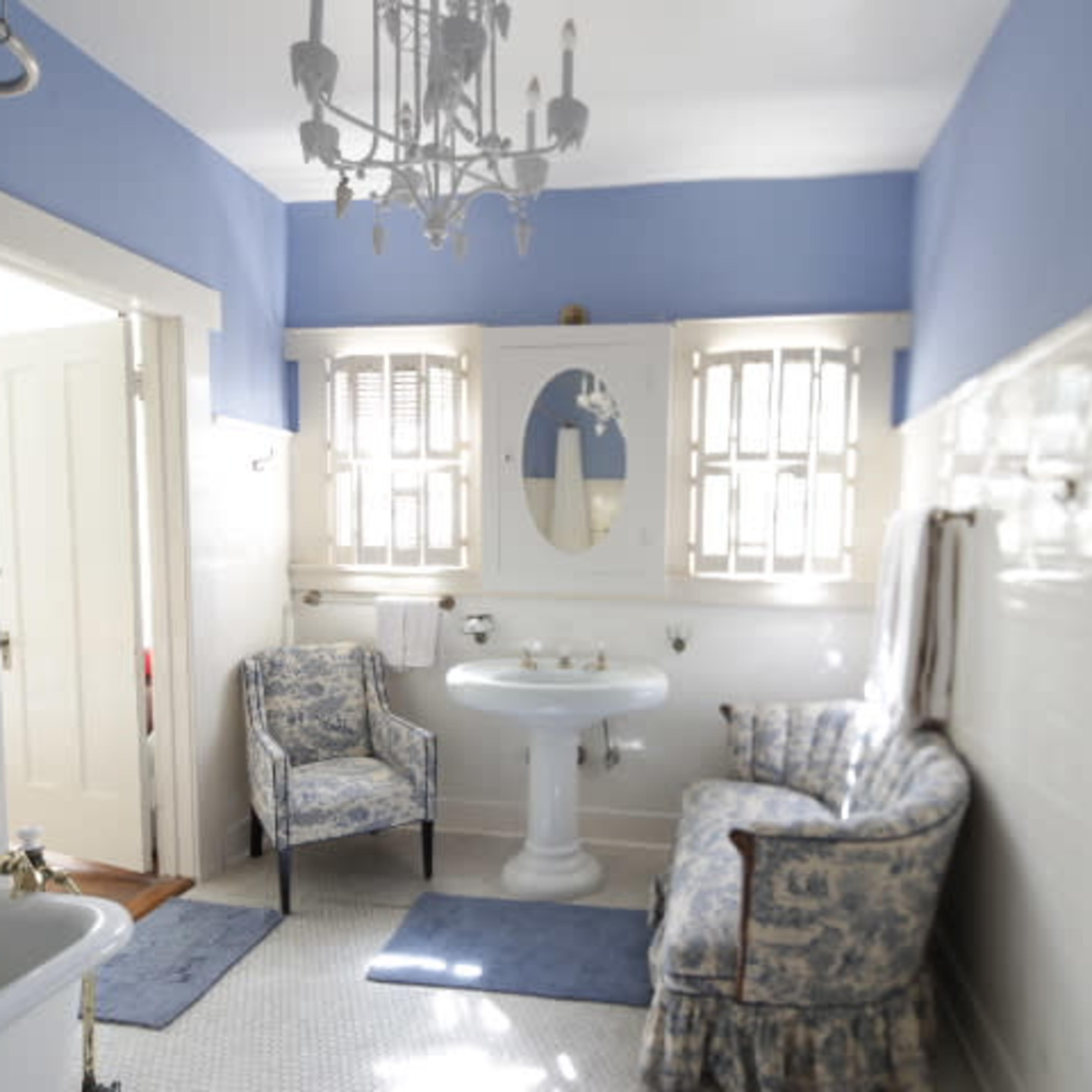 A bright bathroom featuring a clawfoot bathtub, a pedestal sink, two upholstered chairs, and a chandelier, with light blue walls and white tiles.