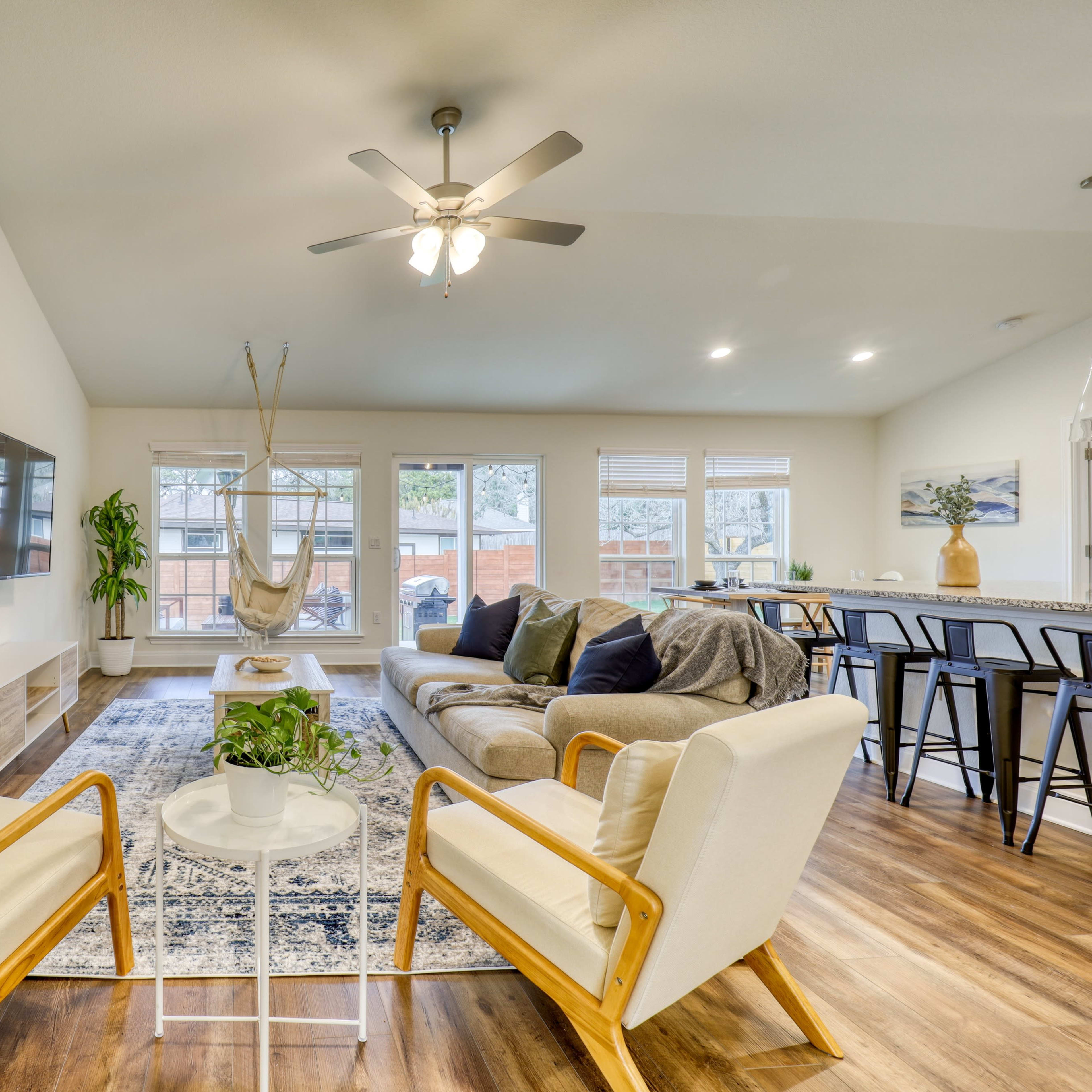 The living room features a neutral color palette with a large sofa, two armchairs, a hanging chair, and large windows providing natural light.