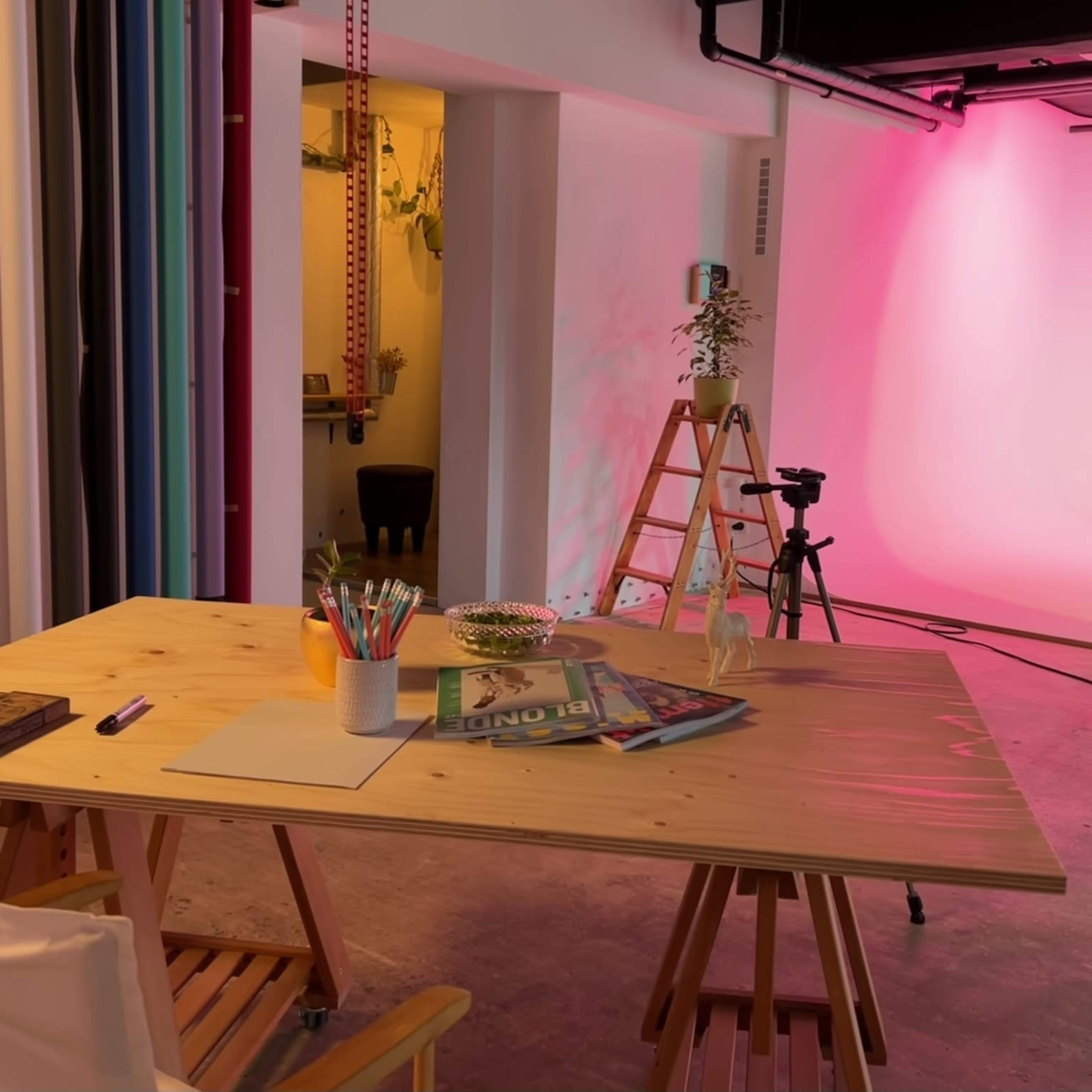 A workspace with a wooden table, colorful art supplies, a camera setup, and a softly lit backdrop in a studio.
