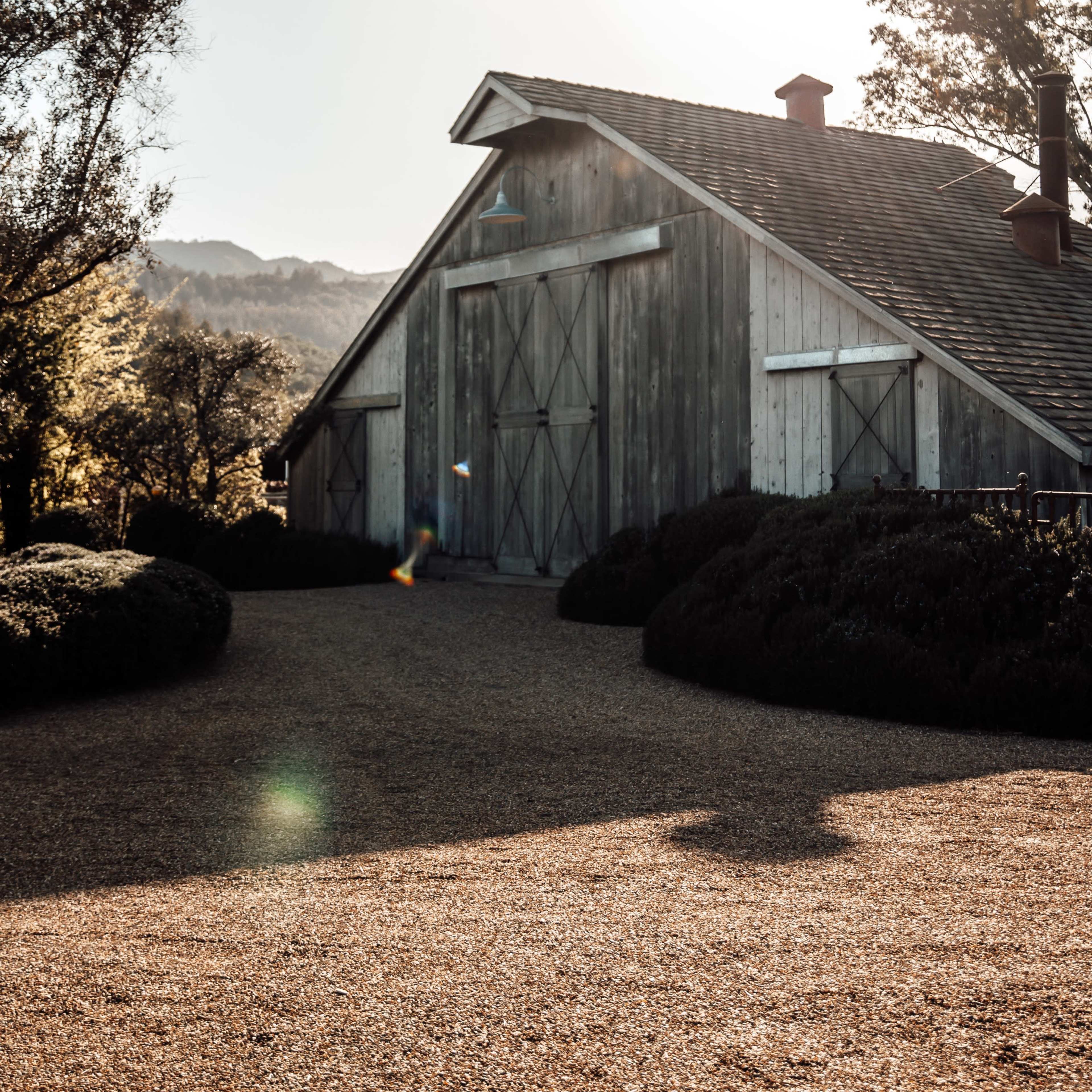 A weathered barn stands beside a gravel driveway, with trees and hills visible in the background under a setting sun.