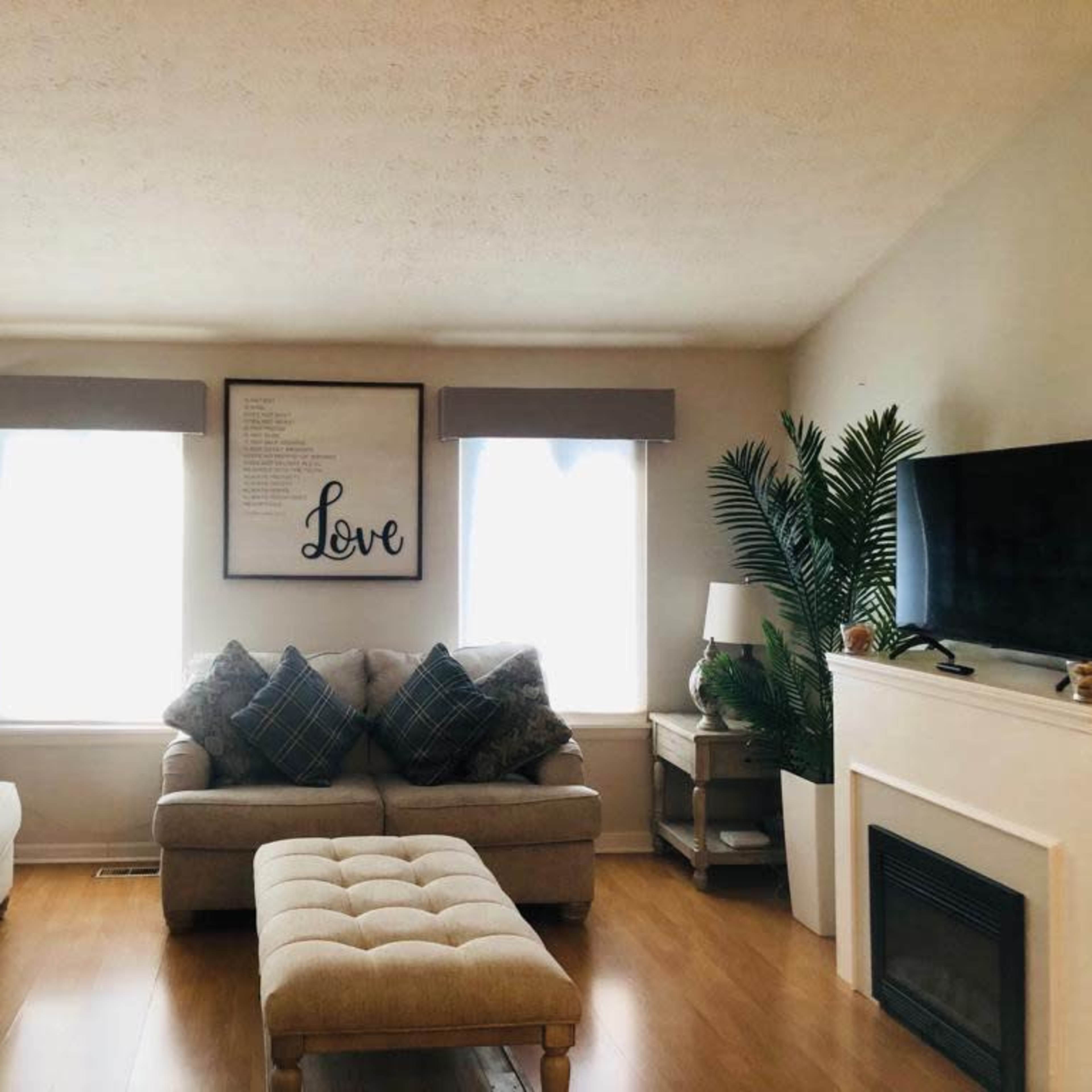 The living room features a beige couch set, two lamps, a large wall art with the word "Love," and a television mounted above a white fireplace.