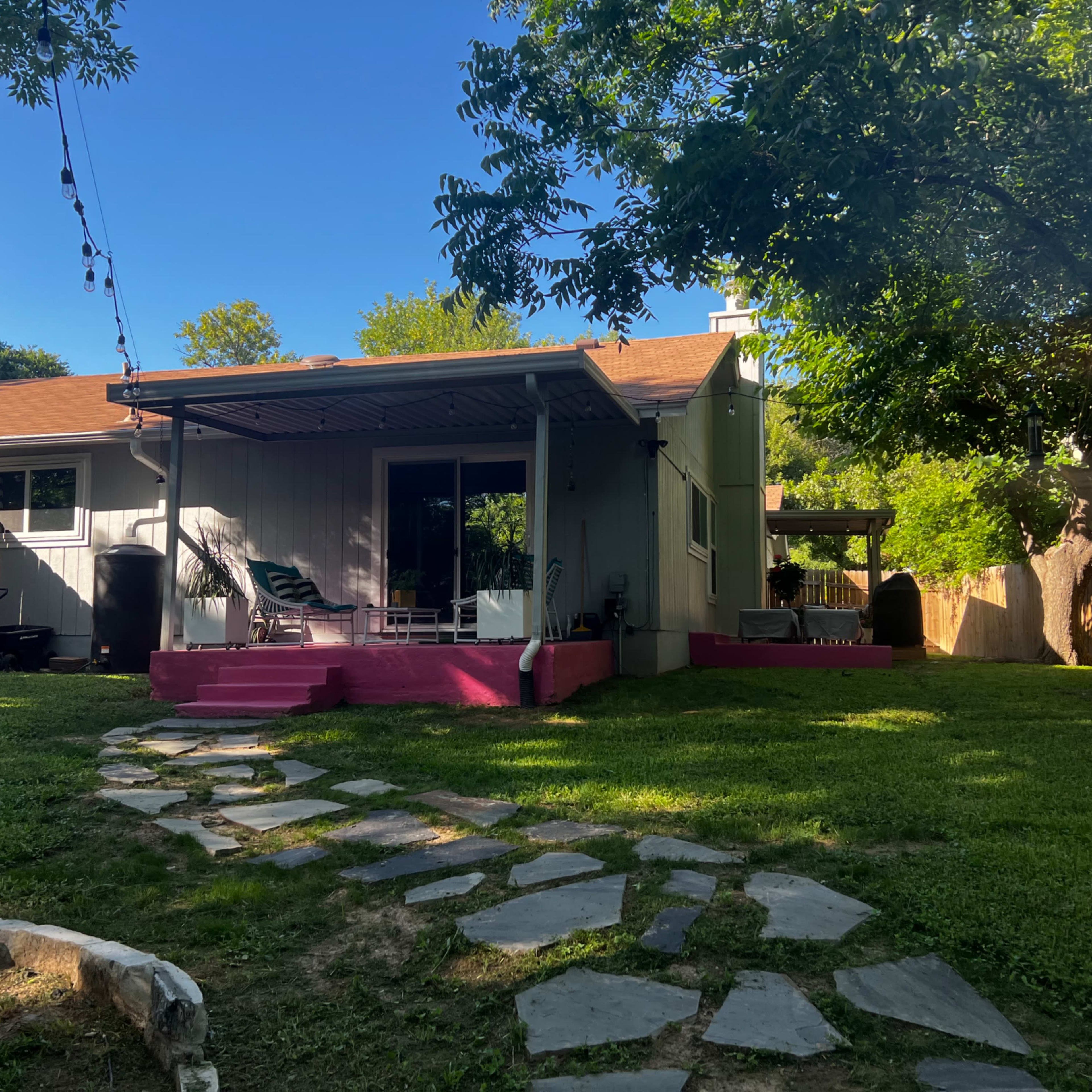 A backyard with a stone path leading to a house featuring a covered patio and a lawn surrounded by trees.