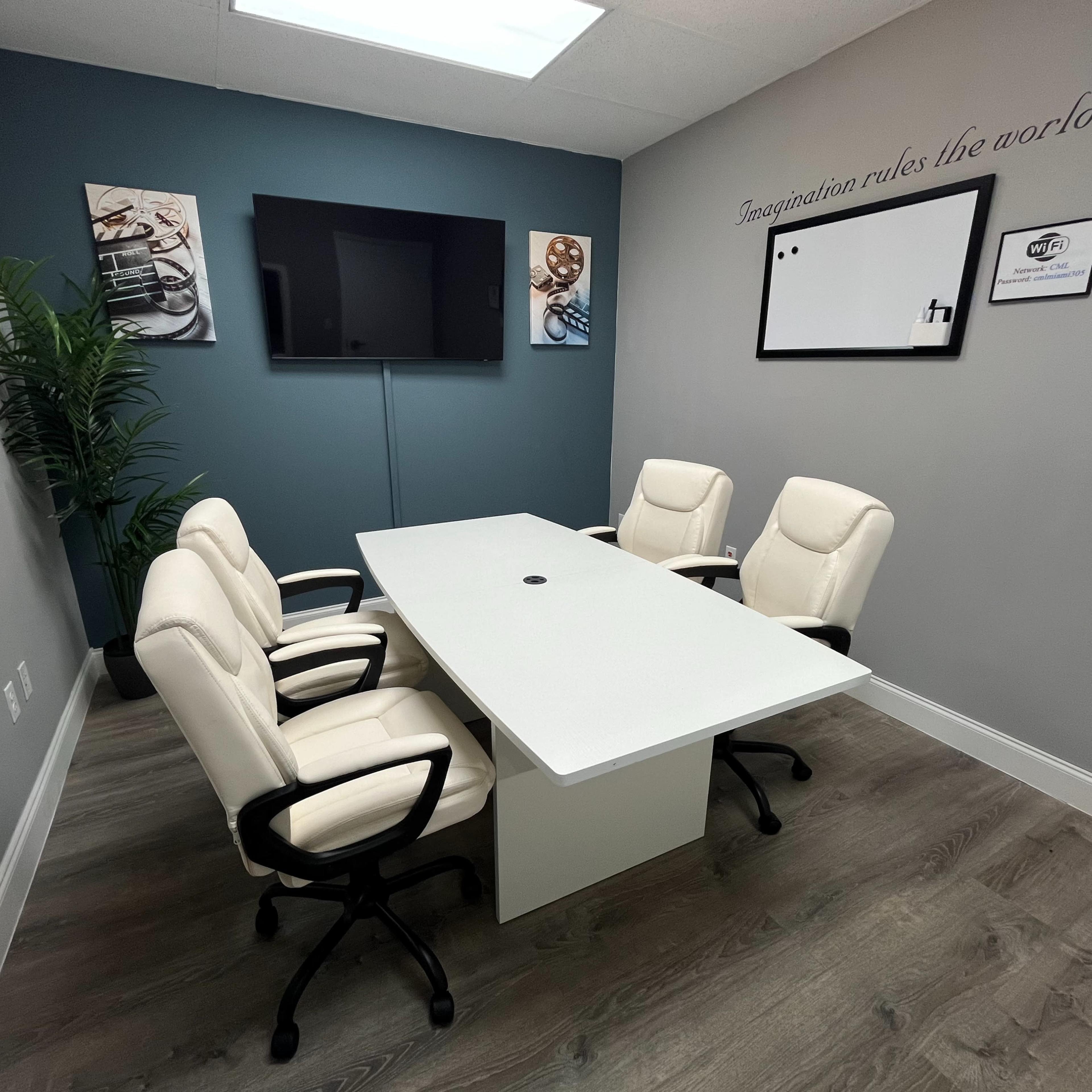 A modern conference room with a rectangular white table surrounded by four white ergonomic chairs, a television mounted on the wall, and motivational artwork.