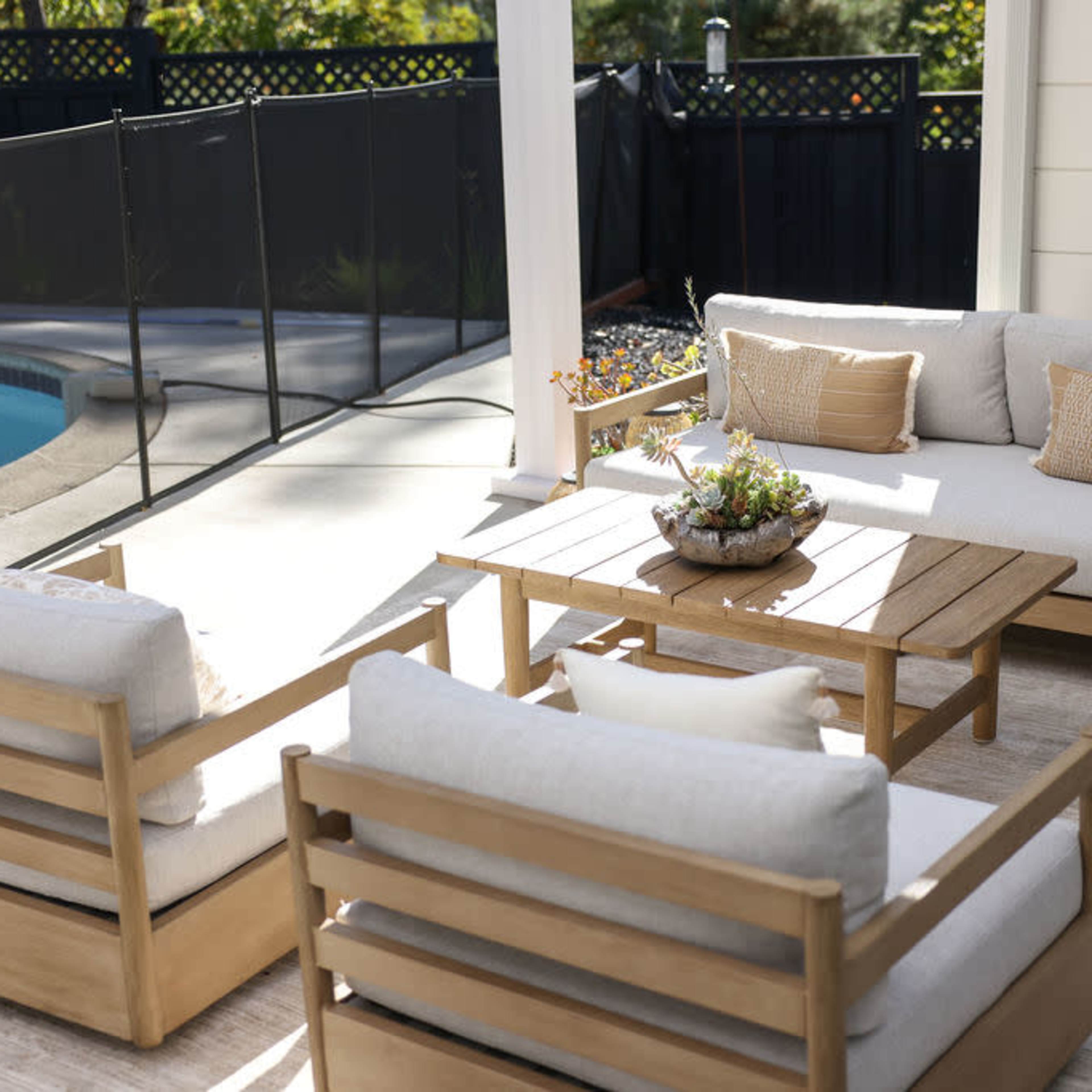 A patio area with a wooden furniture set consisting of a sofa and two chairs around a table, adjacent to a swimming pool.
