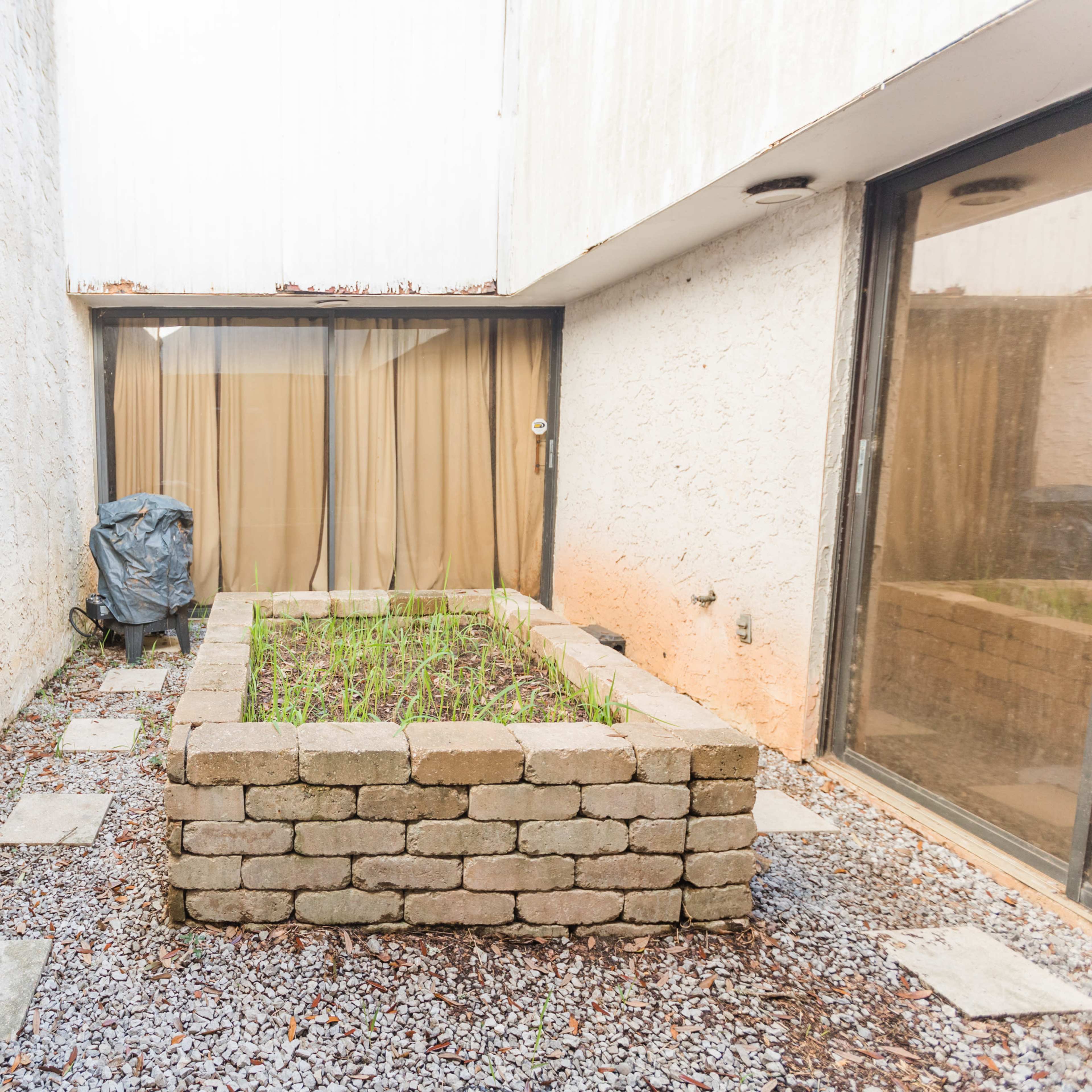 A small courtyard with a raised stone planter bed filled with greenery, surrounded by gravel and concrete walkways.