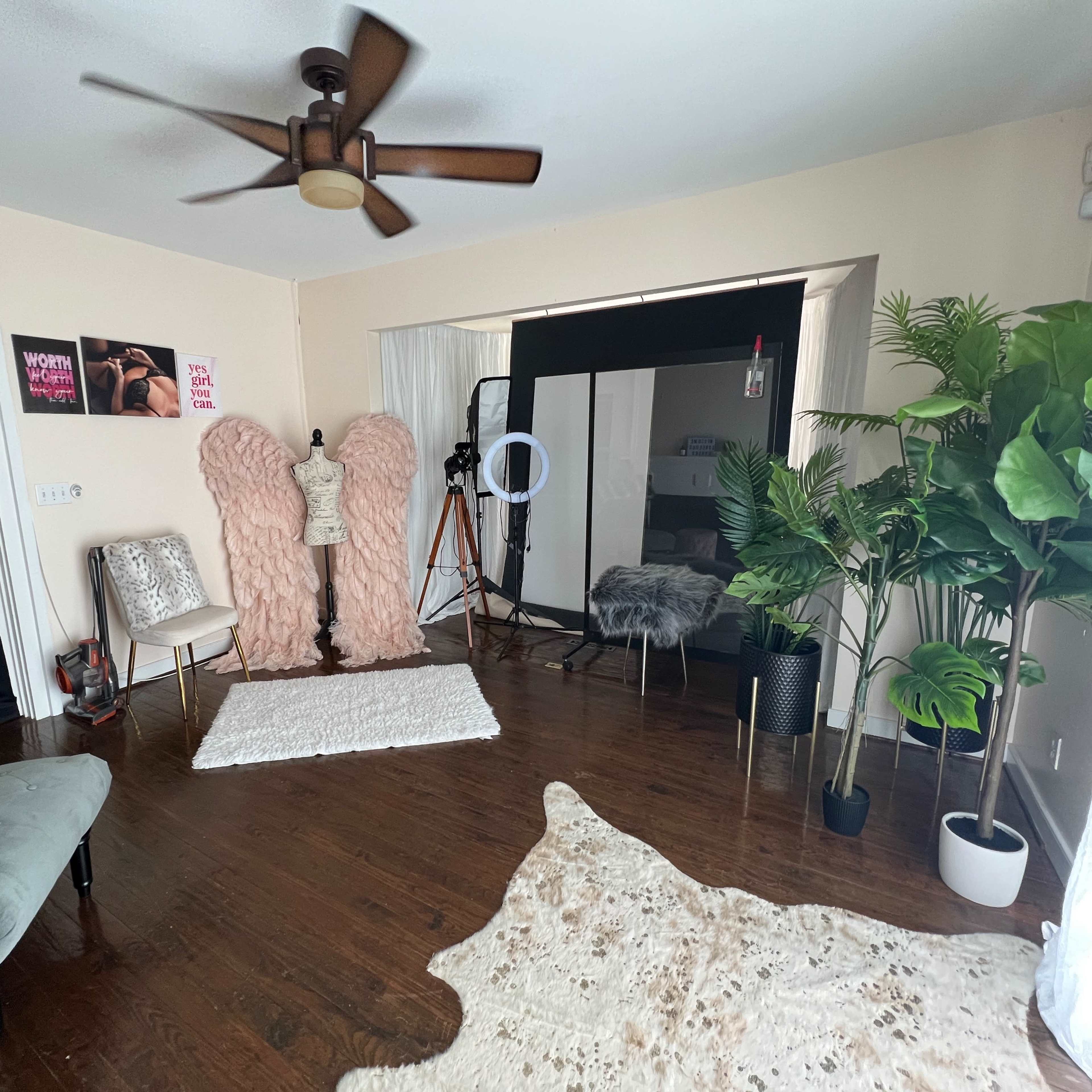 A well-lit room featuring large faux fur angel wings, a clothing rack, a ring light, potted plants, and a soft area rug on a wooden floor.