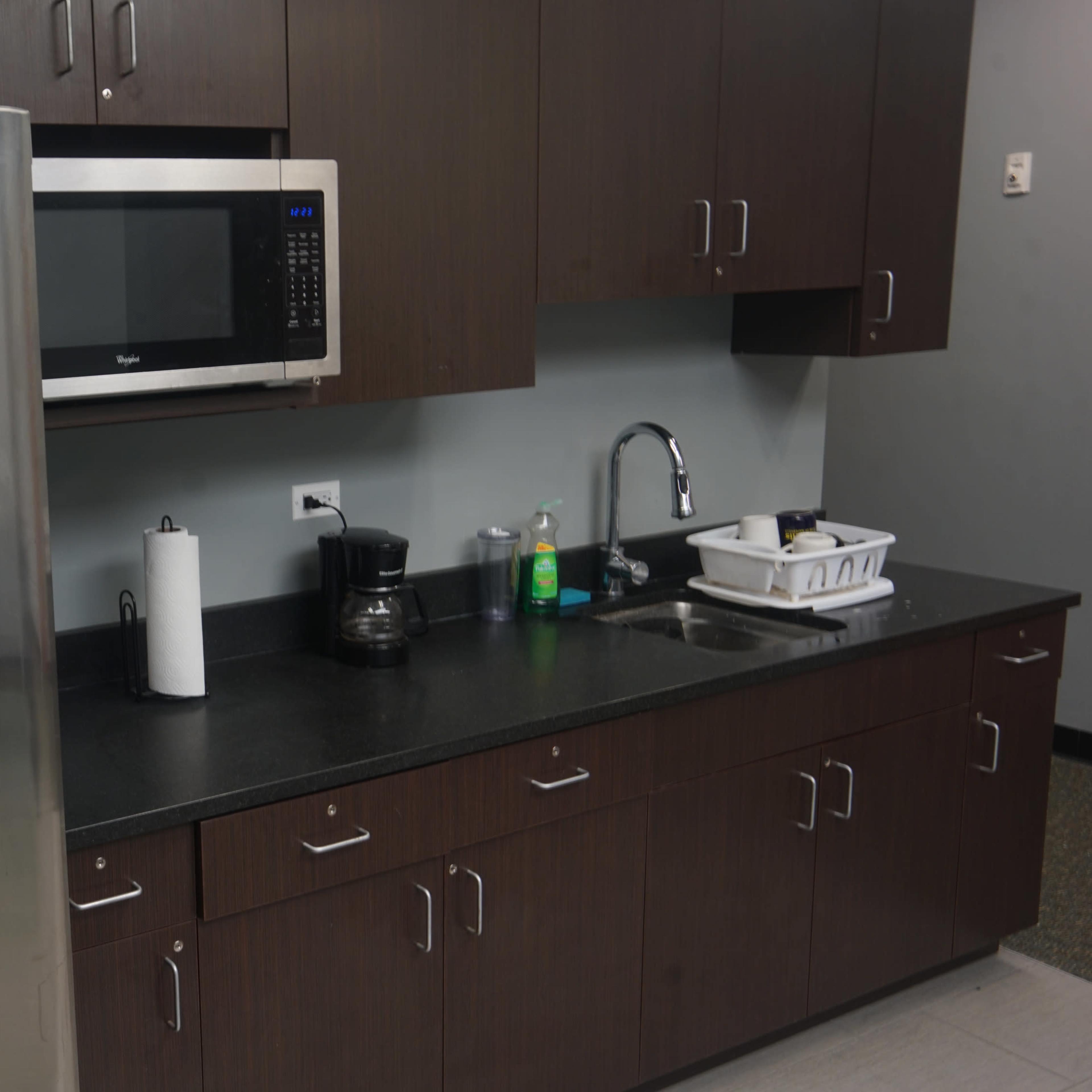 A stainless steel refrigerator and microwave are installed in a kitchen with dark wood cabinets and a black countertop.