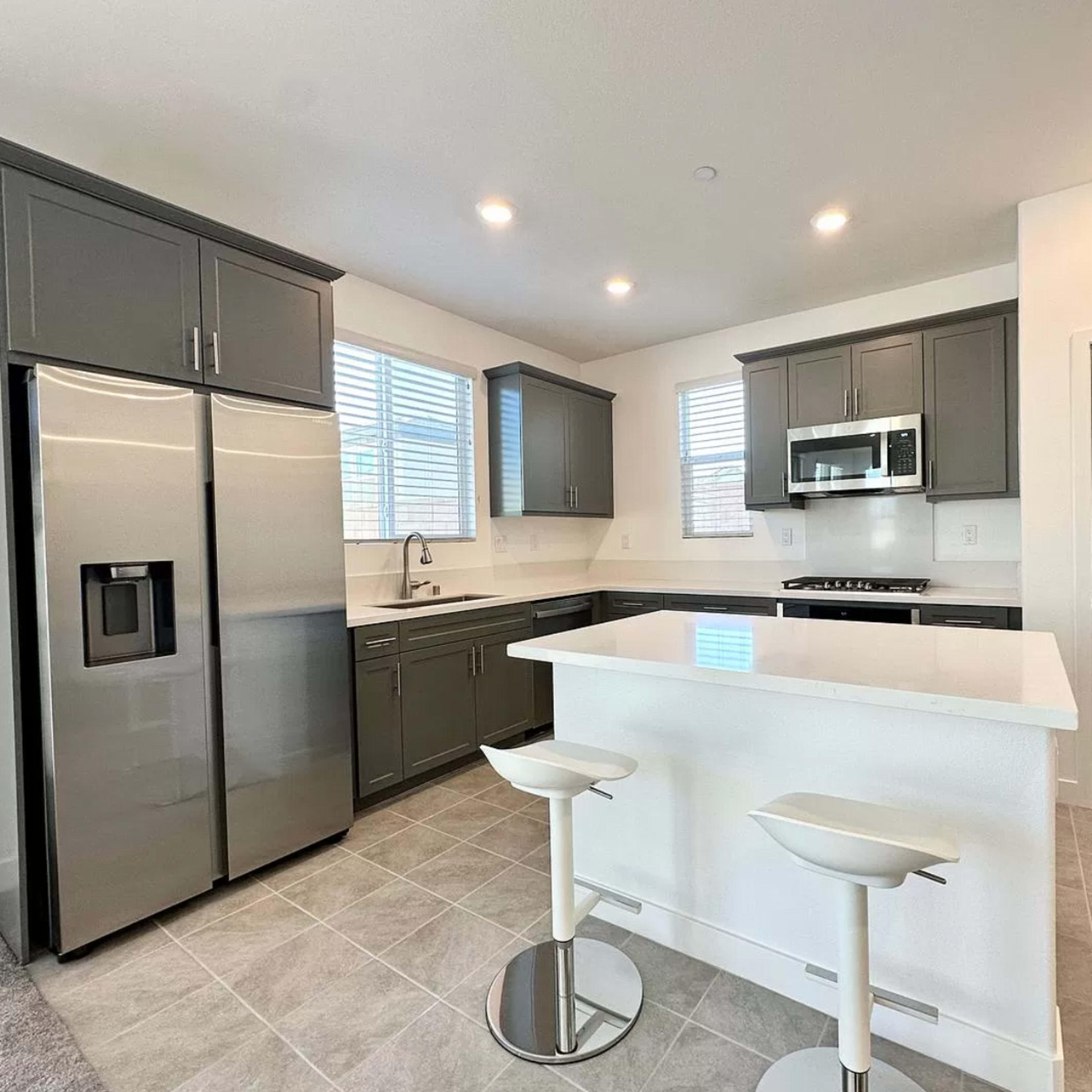 A modern kitchen features gray cabinetry, stainless steel appliances, and a white island with two bar stools.