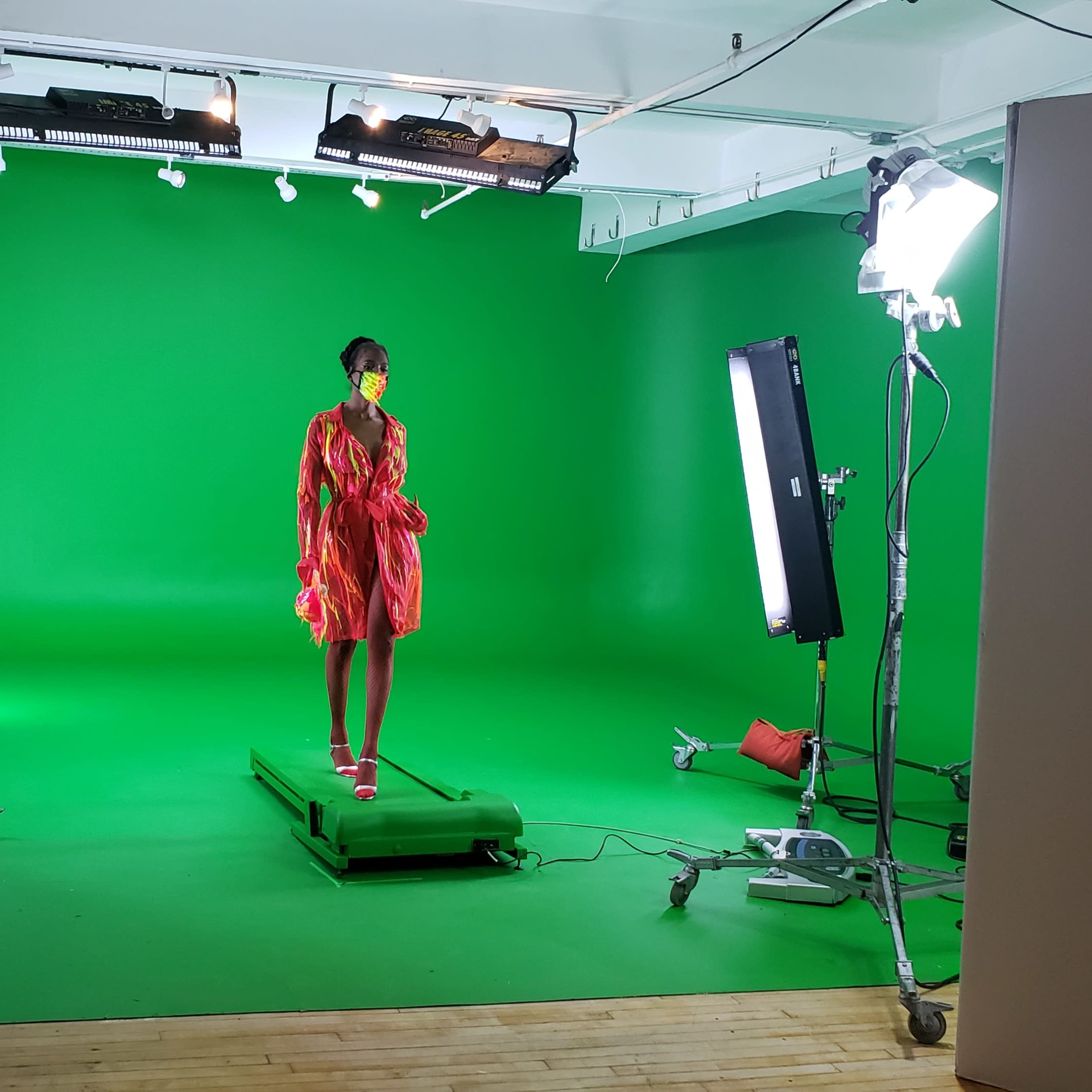 A model stands on a platform in a studio with a green backdrop, surrounded by studio lights and equipment.