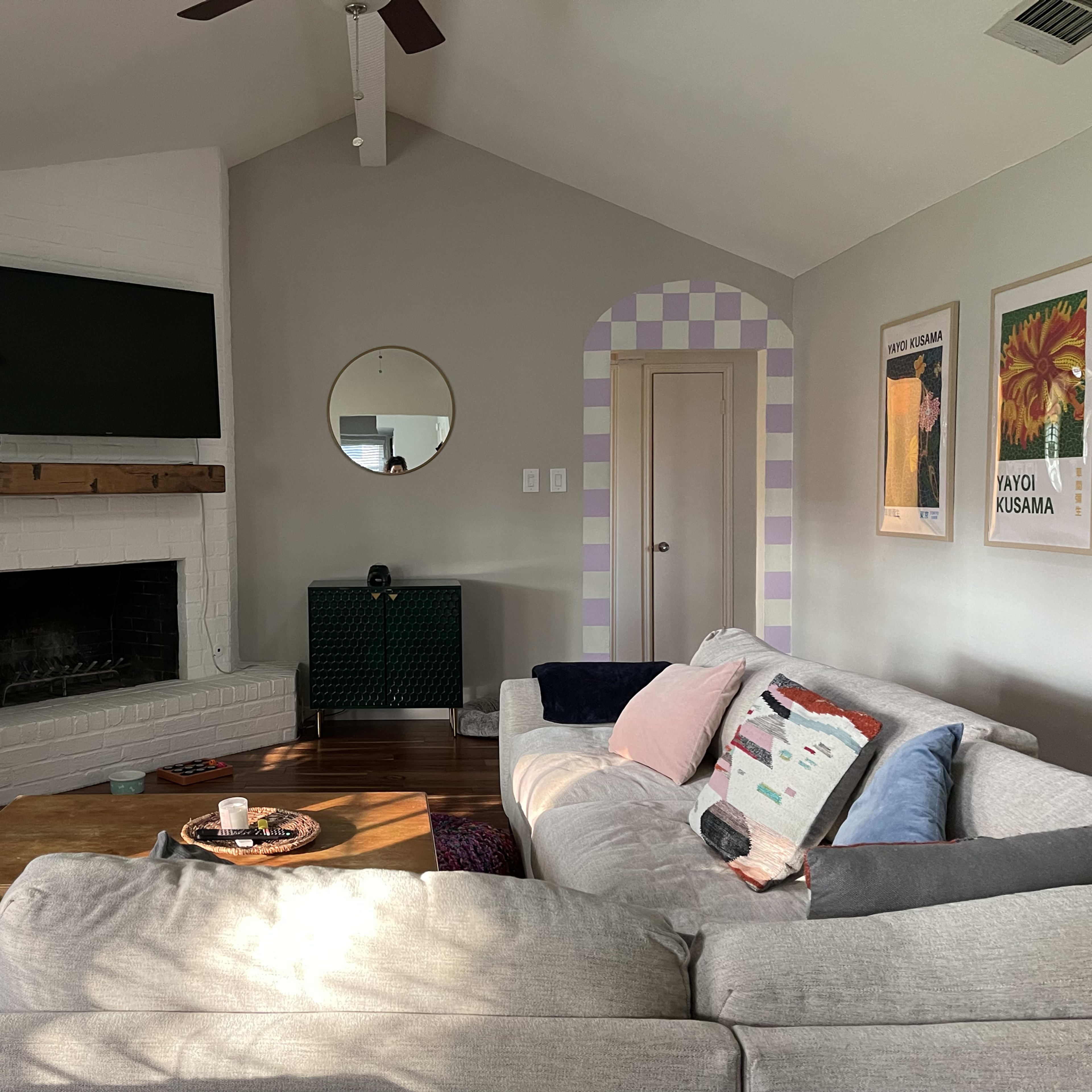 A living room featuring a gray sectional sofa, a white brick fireplace with a mounted TV, and colorful artwork on the wall.
