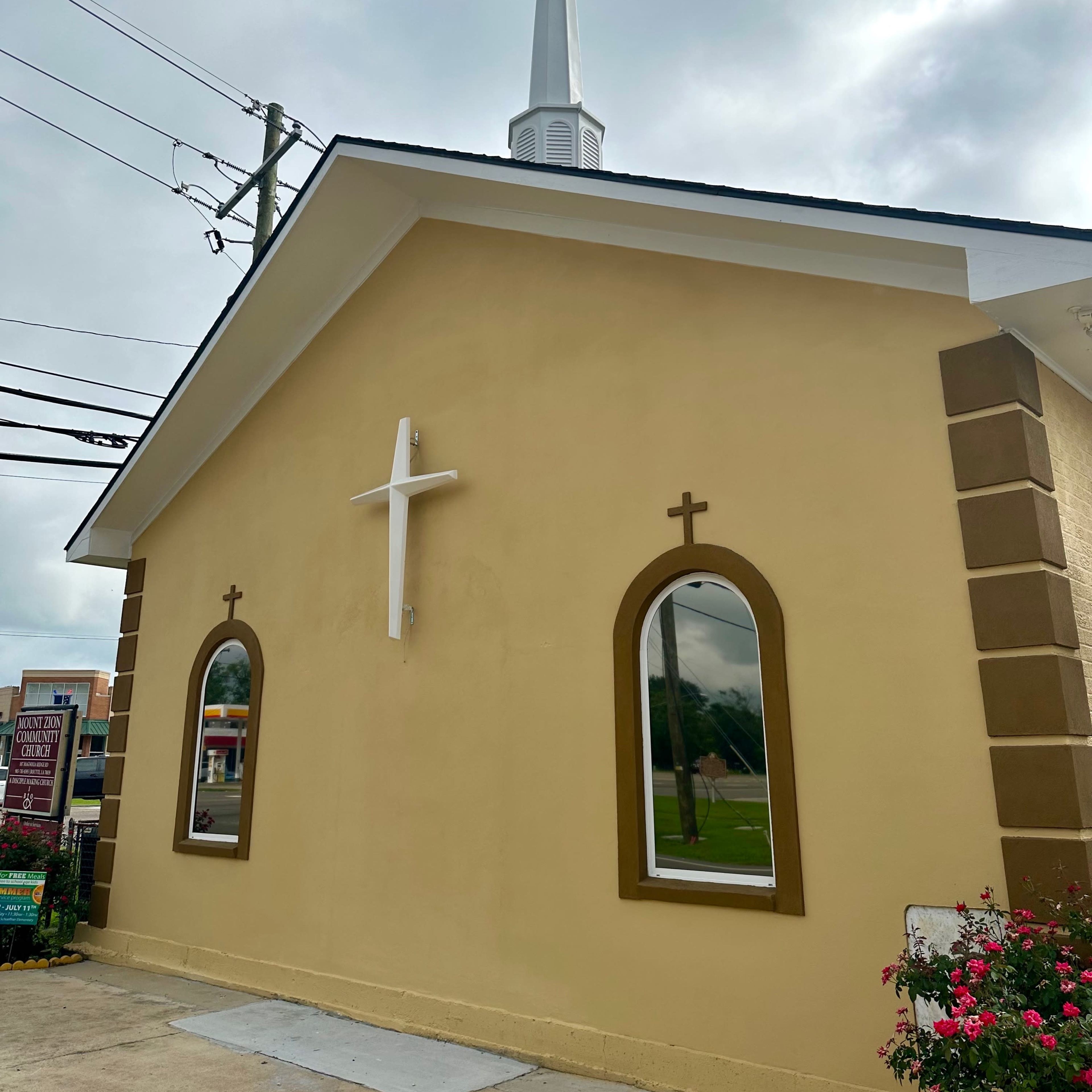 The side of a church building featuring a tall white steeple, two arched windows with crosses, and a prominent white cross on the wall.