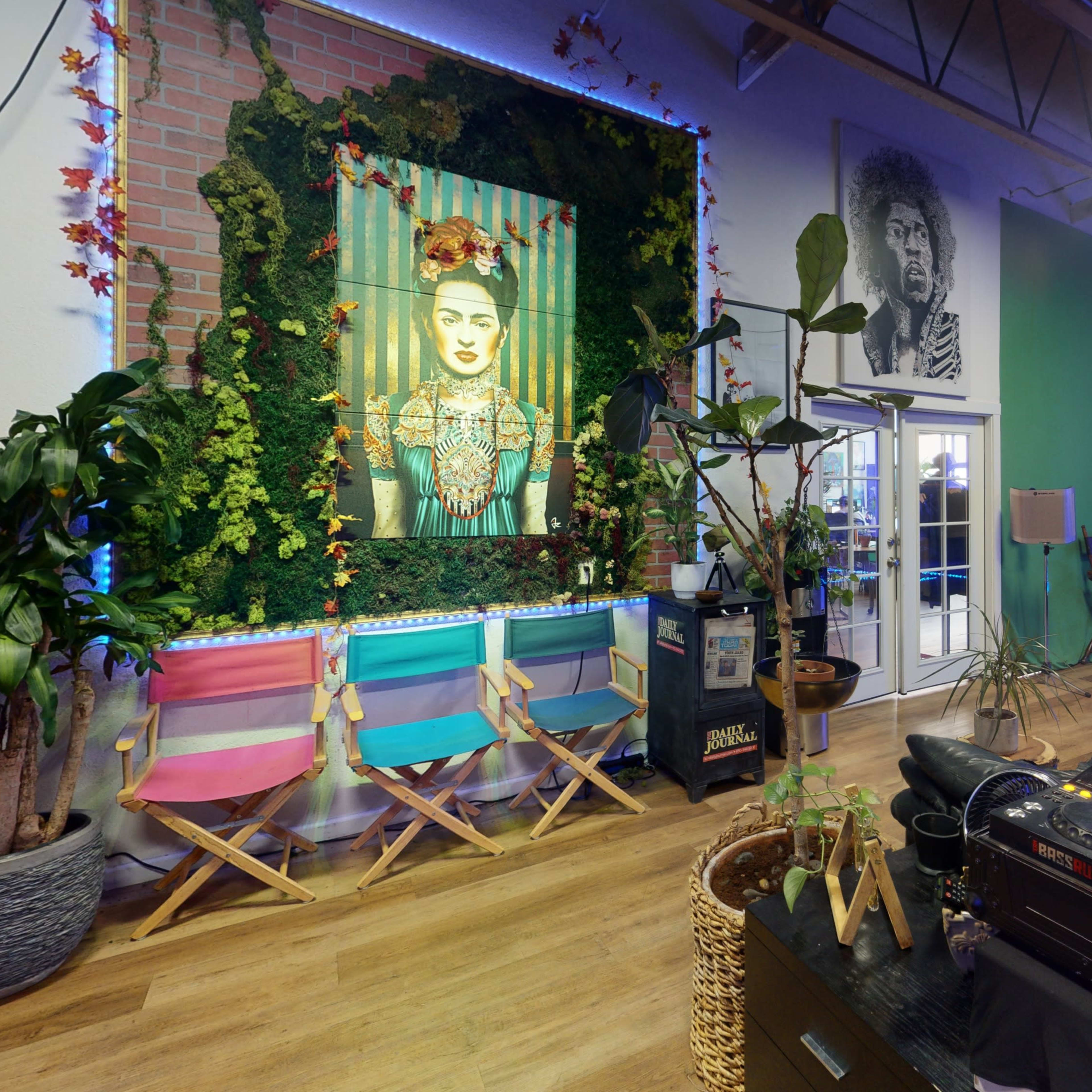 A vibrant creative space featuring a large portrait of Frida Kahlo, a performing stage, colorful chairs, and a DJ setup with plants decoratively arranged throughout.