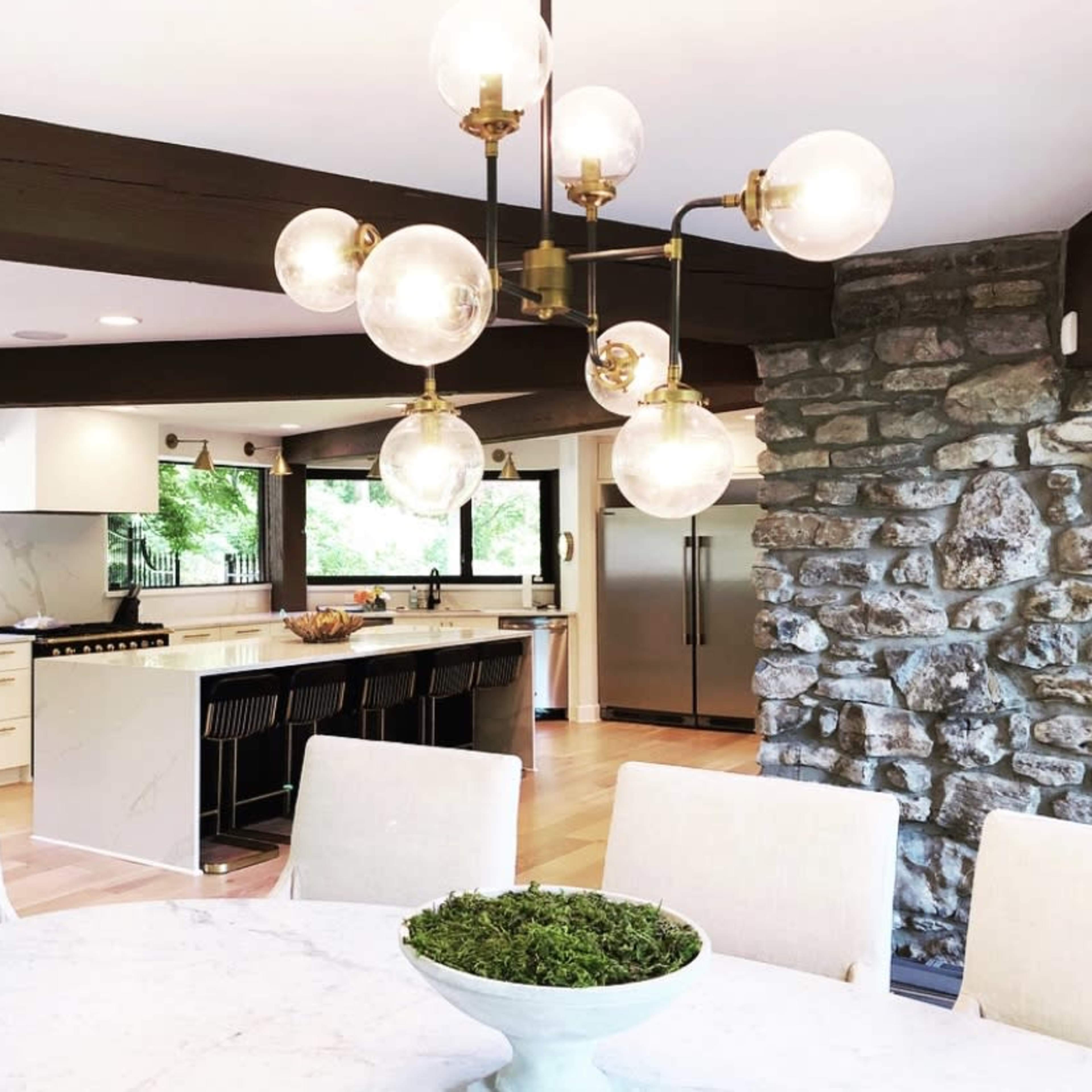 A modern kitchen features a round dining table with a green centerpiece, a stone wall, and a stylish chandelier with round glass bulbs.