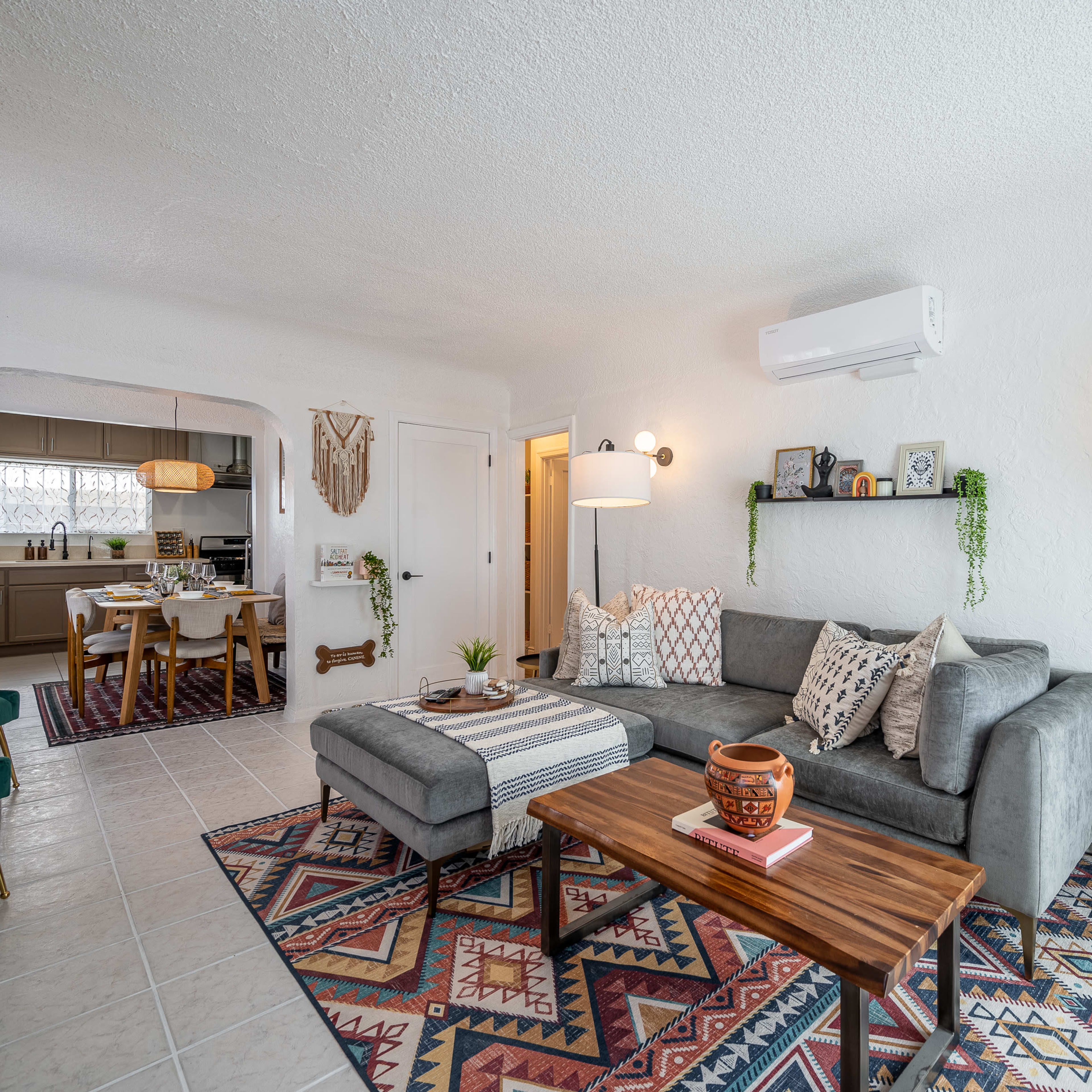 A modern living room features a gray sectional sofa, a wooden coffee table, and vibrant rugs, with a dining area visible in the background.