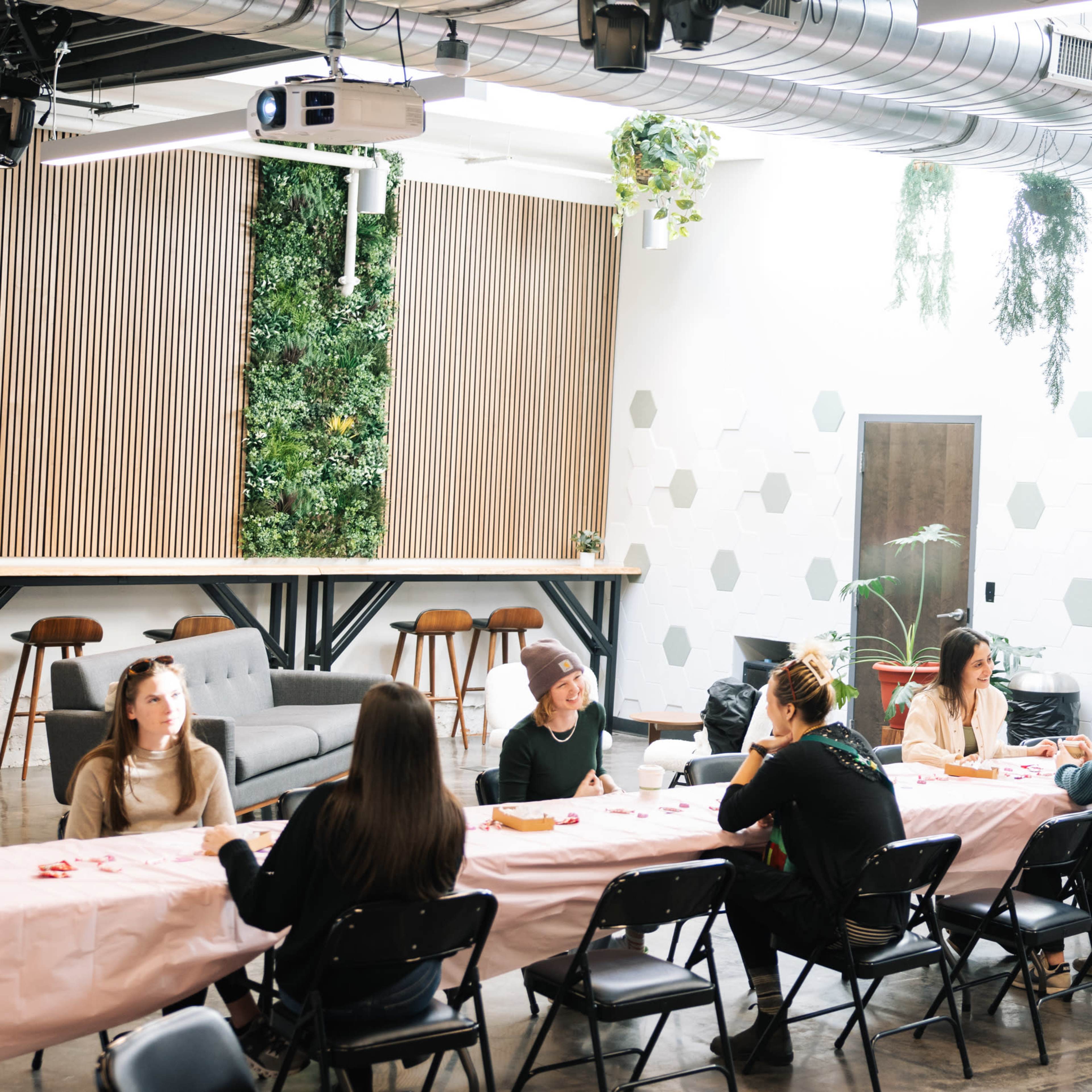 A group of people sits around a long table with a pink tablecloth in a modern meeting space featuring green wall decor and overhead plants.