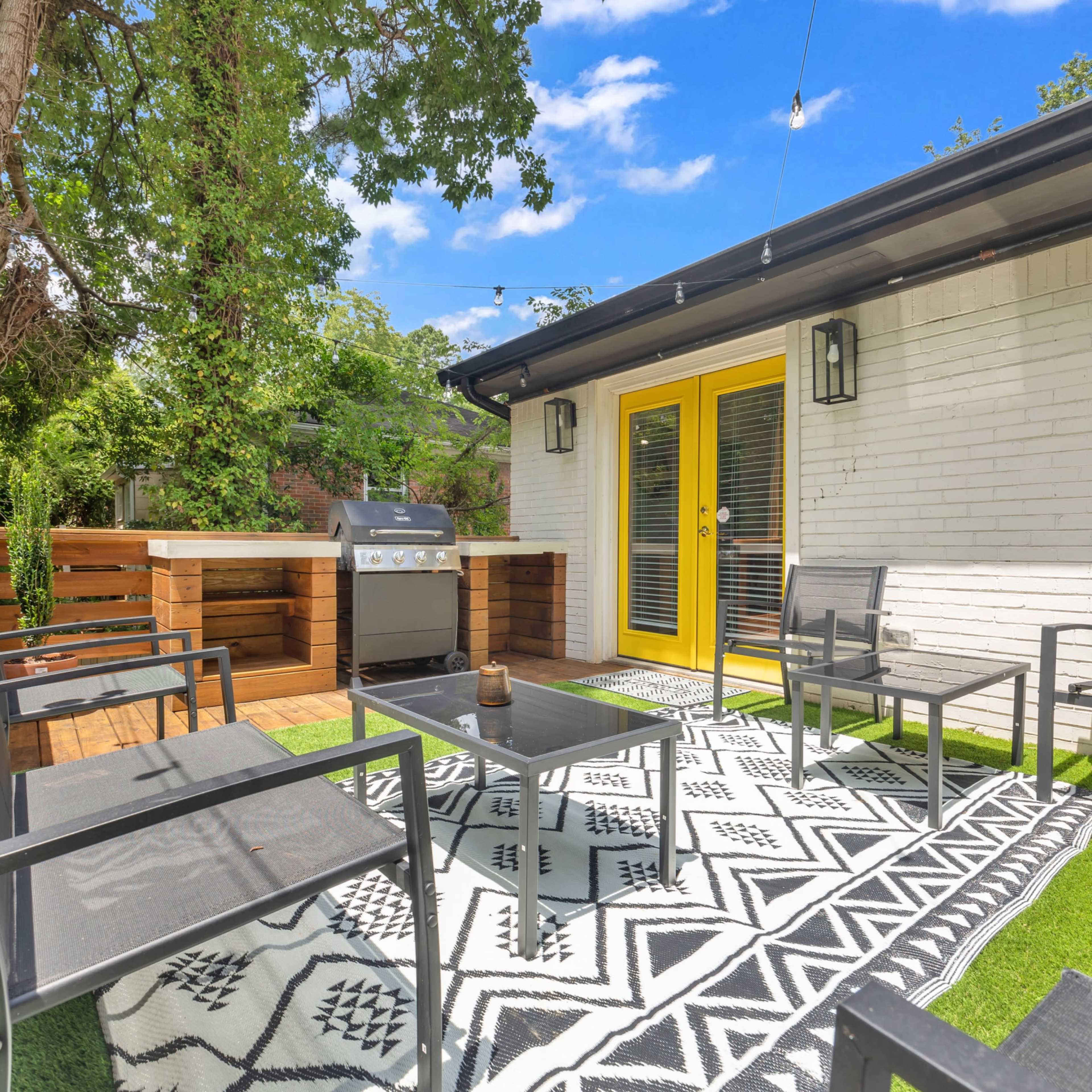 A patio area features a grill, seating with chairs and a table, and is surrounded by greenery with bright yellow doors.