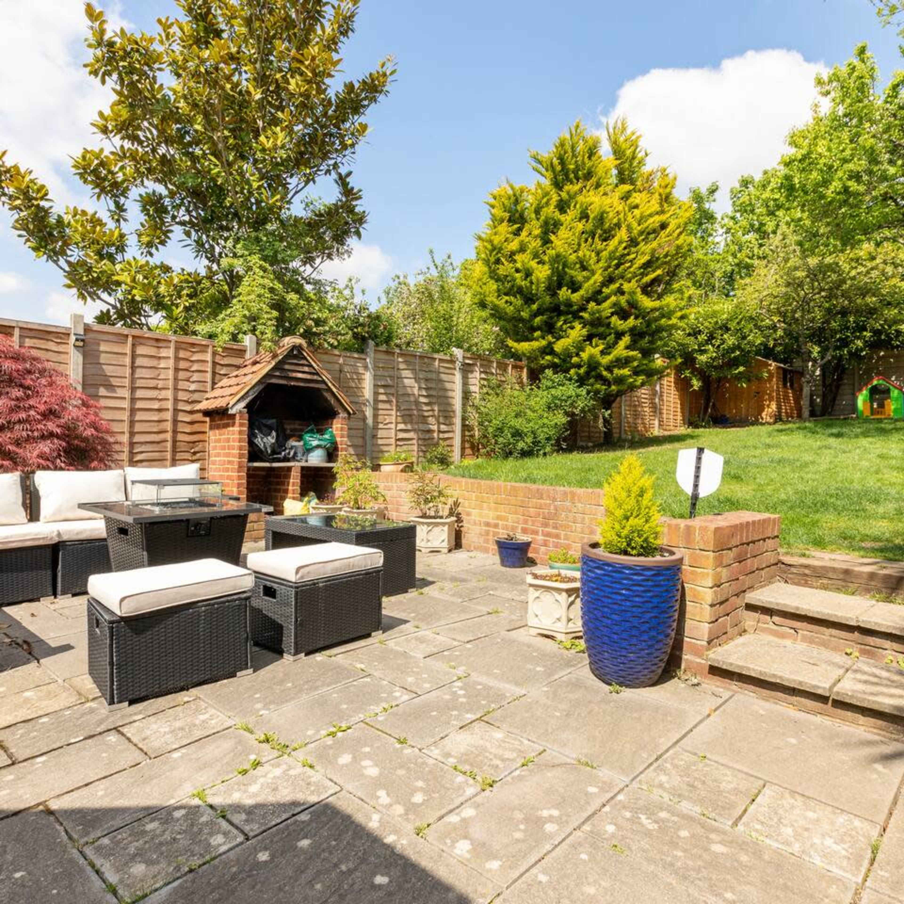 A well-maintained garden with a patio area featuring outdoor furniture, a wooden playhouse, and a grassy space surrounded by trees and shrubs.