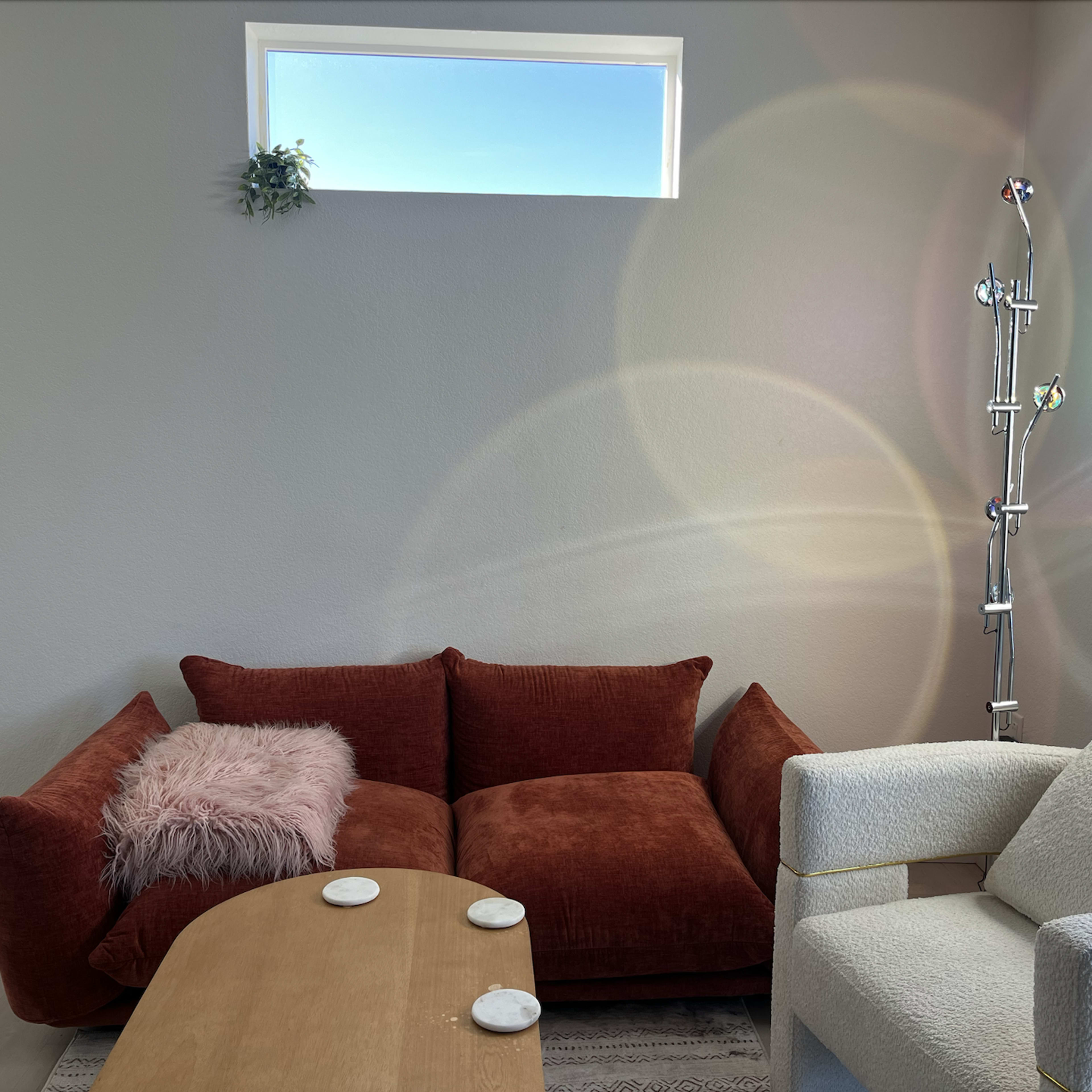 A cozy living room with a brown couch, a white armchair, a wooden coffee table, and a small window allowing sunlight to enter.