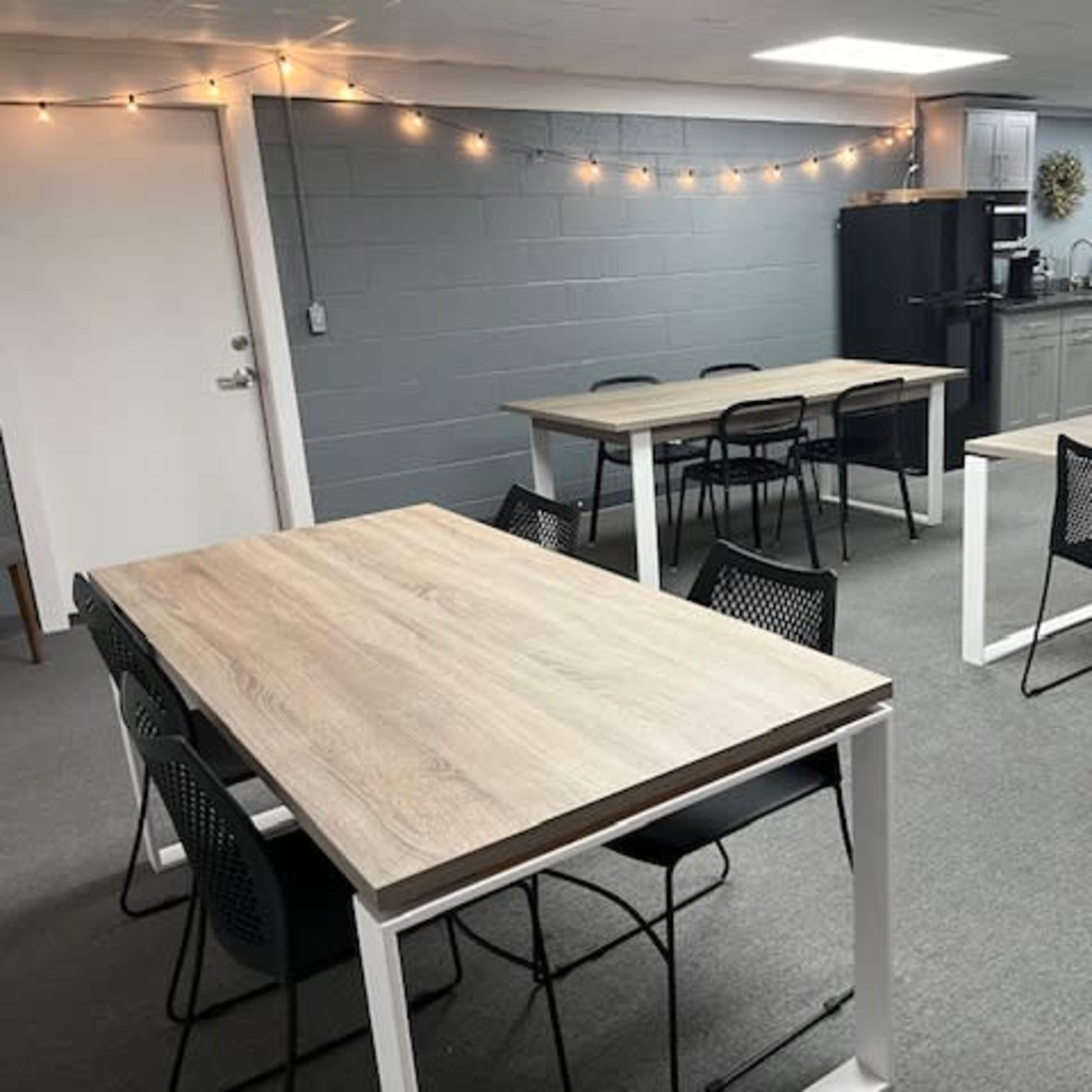 A room with several wooden tables and chairs arranged, along with a kitchenette area featuring a black refrigerator and light fixtures along the wall.