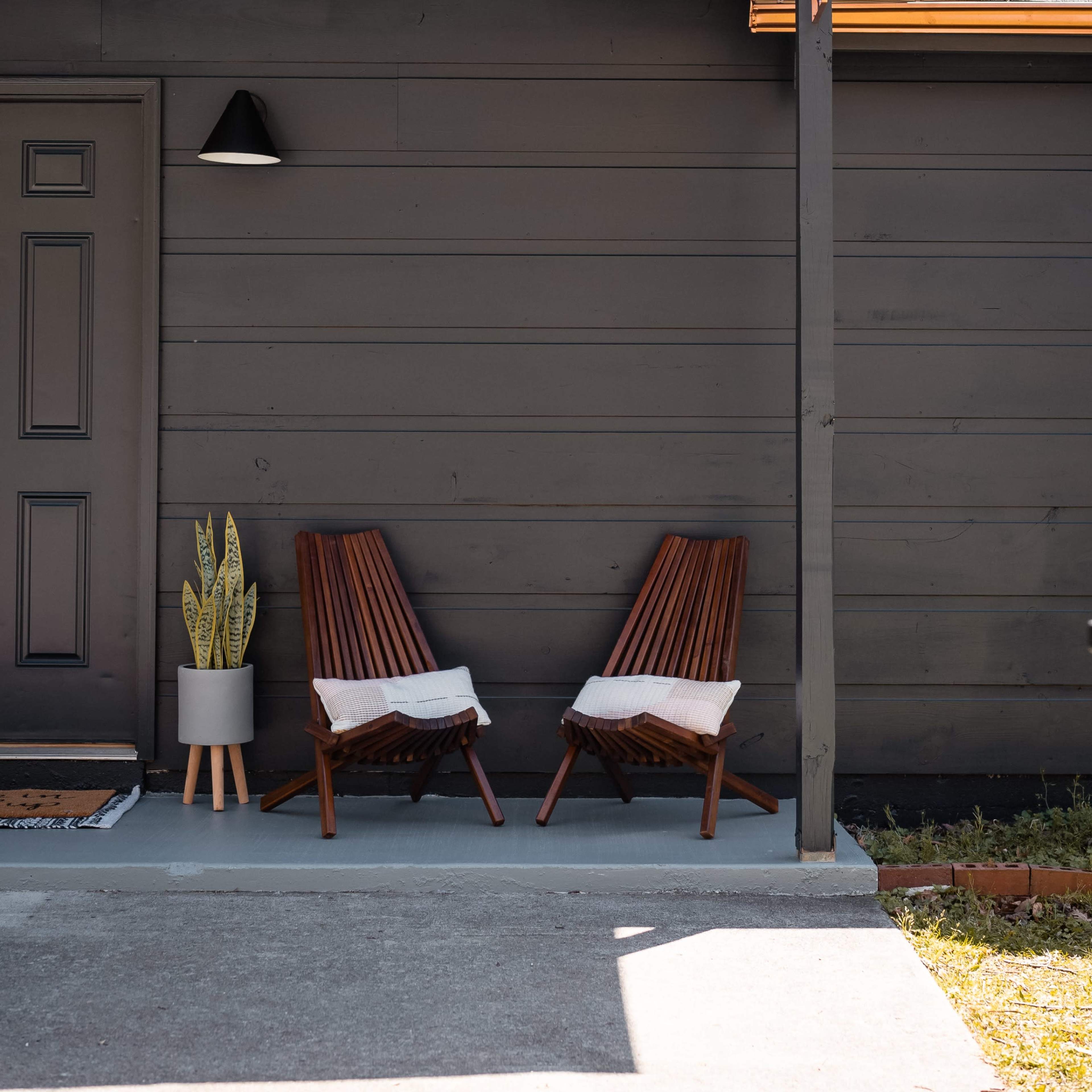 Two wooden lounge chairs with cushions are positioned on a concrete patio next to a potted plant against a dark-colored house exterior.