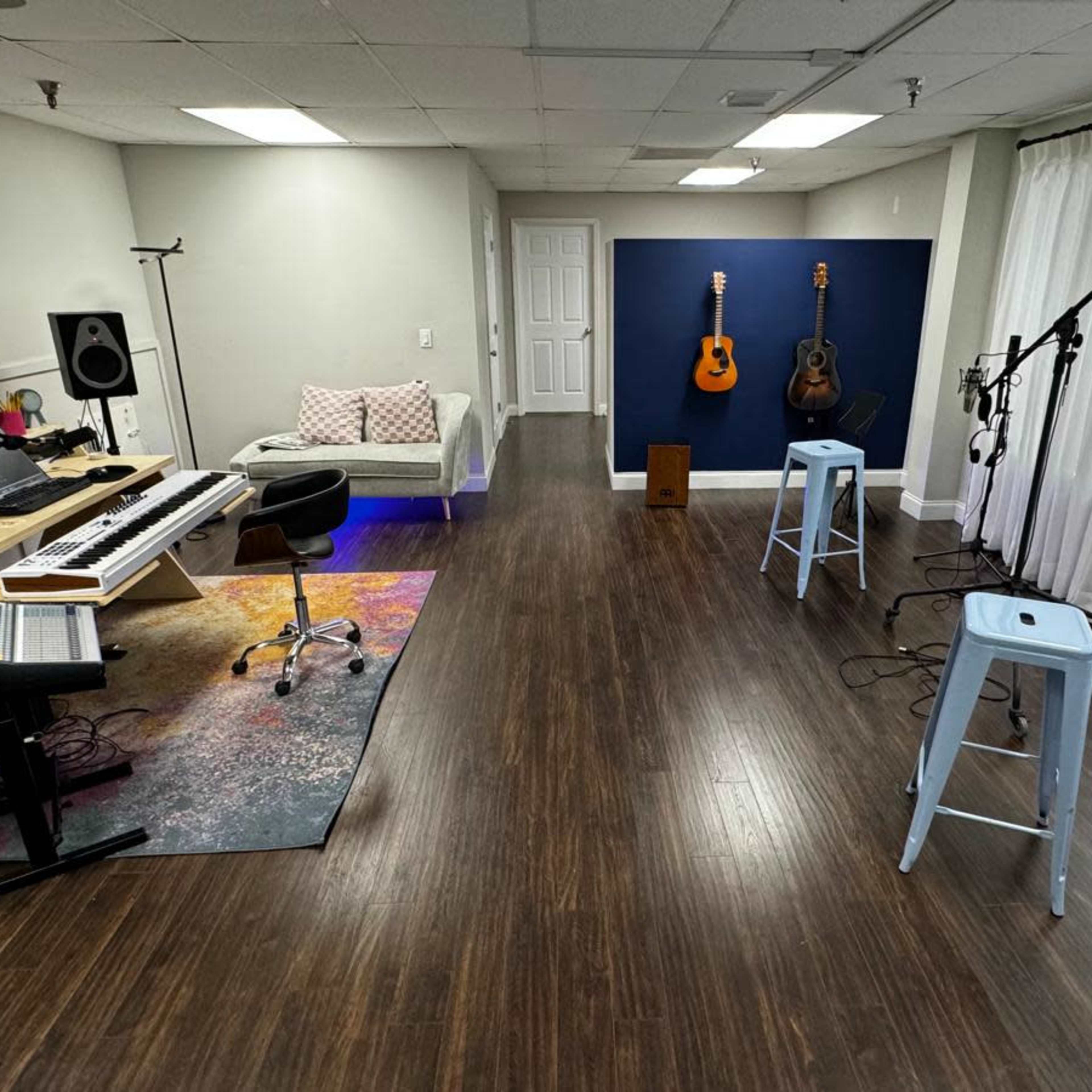 A modern music studio with a keyboard, sound equipment, and instruments, featuring a seating area and large windows.
