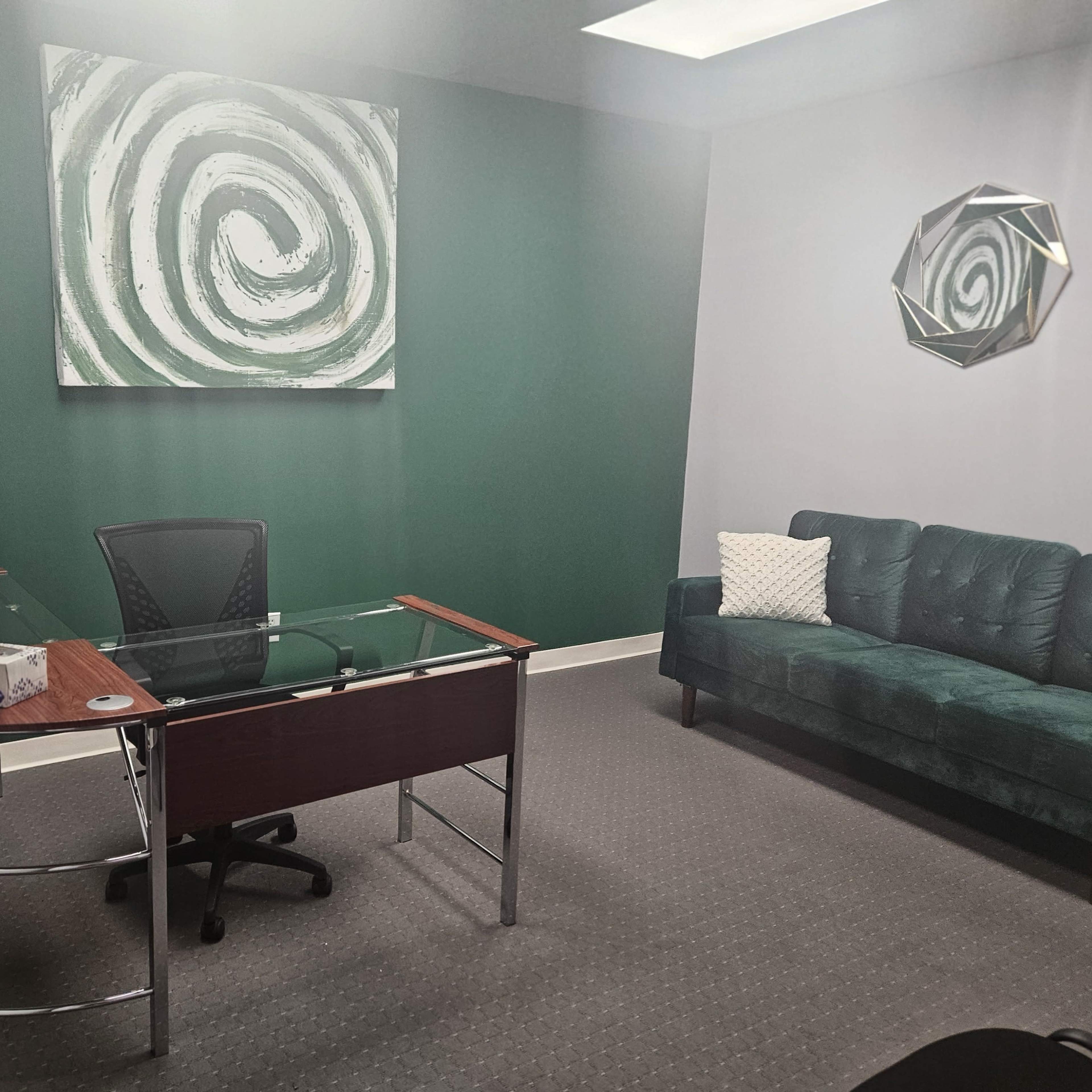 An office space featuring a glass-topped desk, a black office chair, a green accent wall with abstract art, and a green velvet couch.
