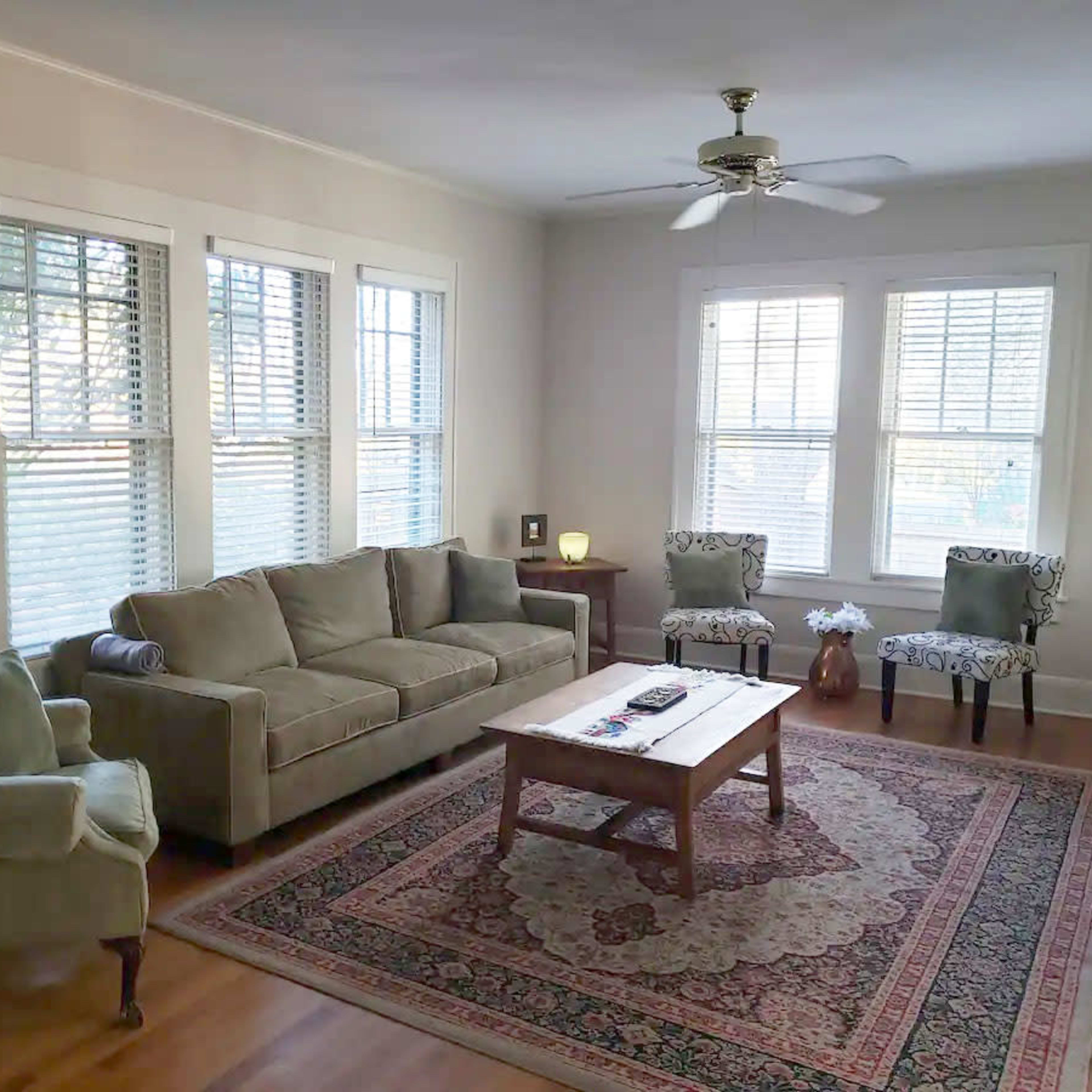 The room features a green sofa, two patterned chairs, a wooden coffee table, and large windows with blinds.