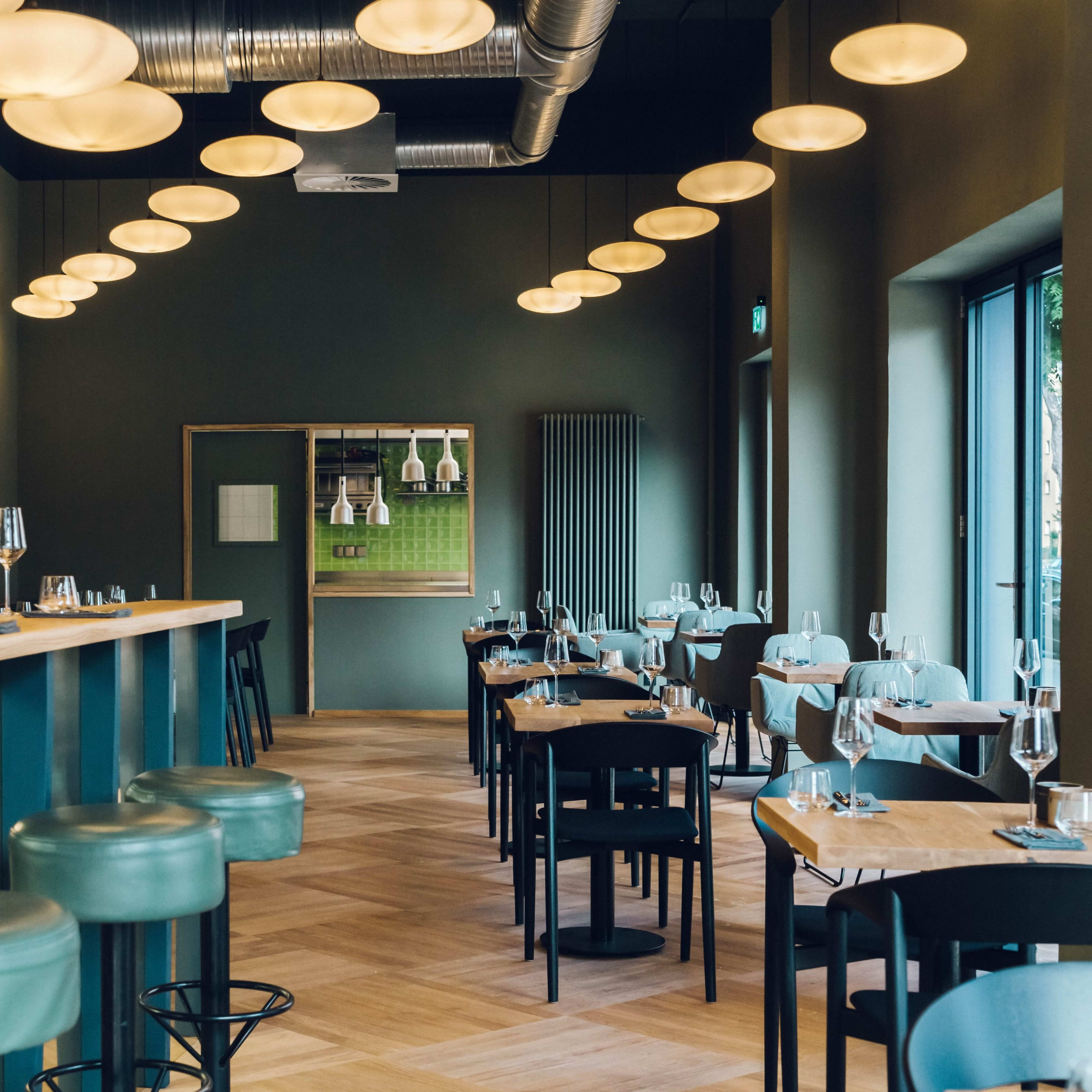 A modern restaurant interior with green walls, wooden flooring, and several round pendant lights hanging from the ceiling, along with spaced-out tables and bar seating.