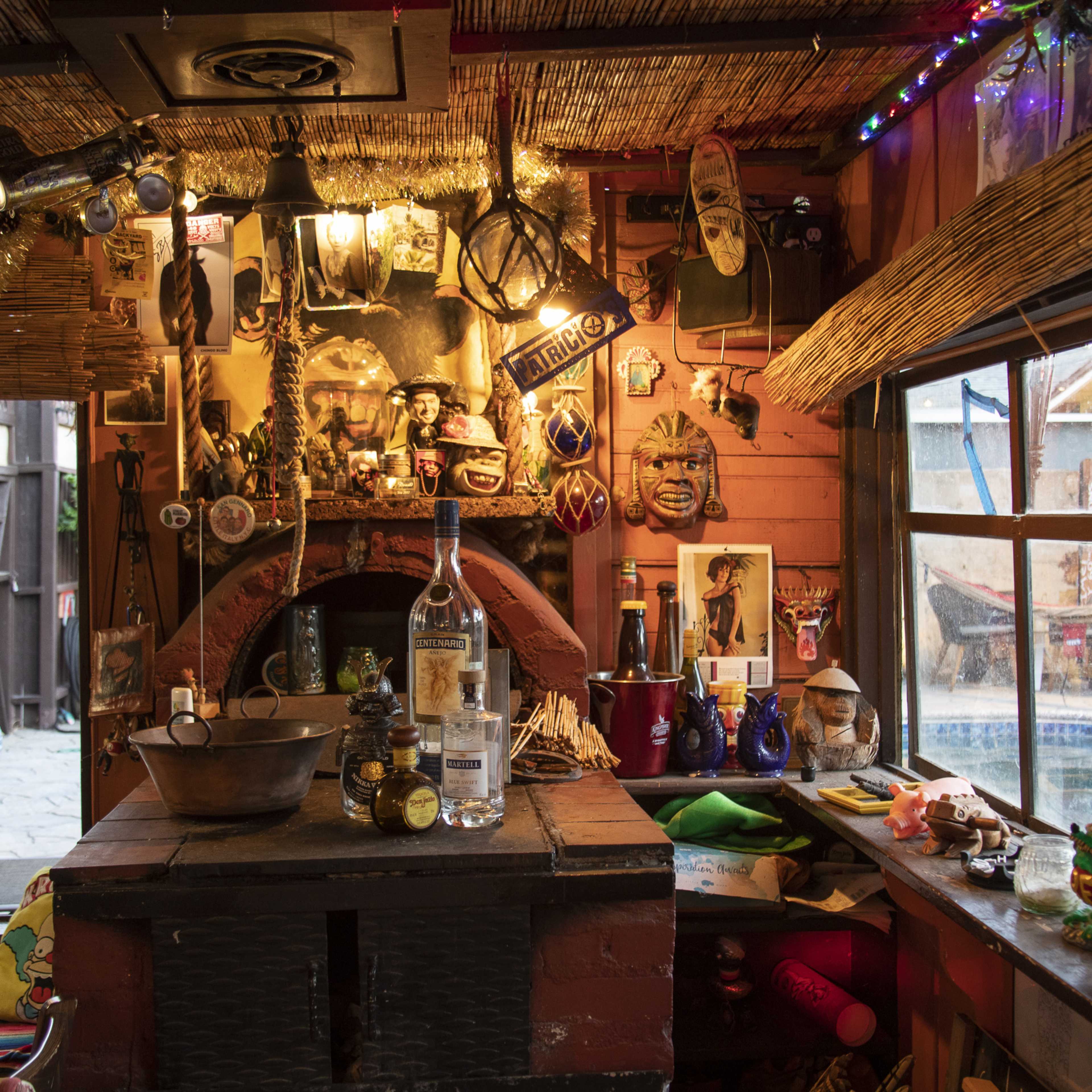 A vibrant indoor bar area decorated with various artifacts, masks, and colorful lights, featuring a central brick oven and shelves stocked with bottles.