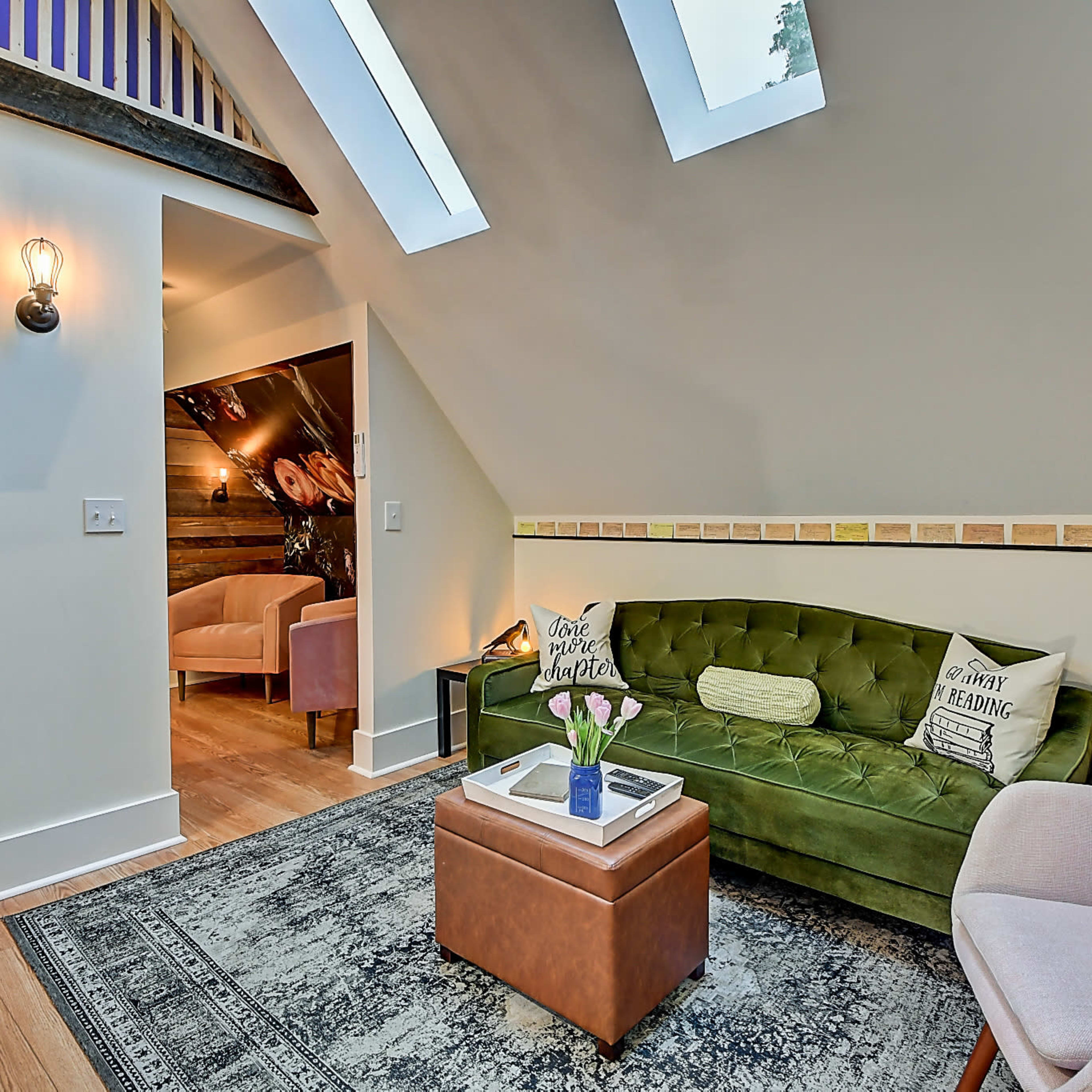 A cozy living space featuring a green sofa, a coffee table, and a pink chair, with skylights and a decorative wall.