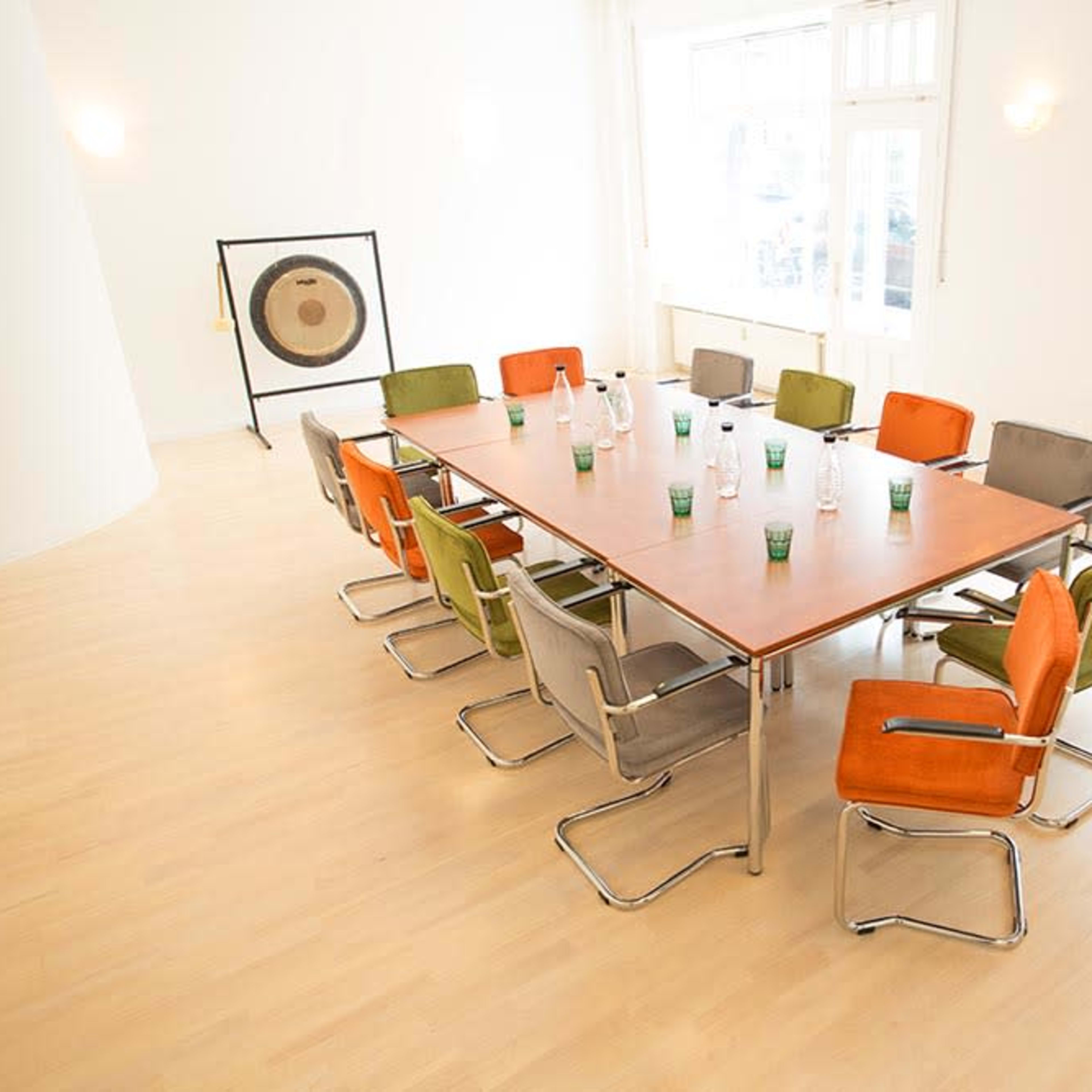 A spacious meeting room features a large rectangular table surrounded by colorful chairs and water bottles.
