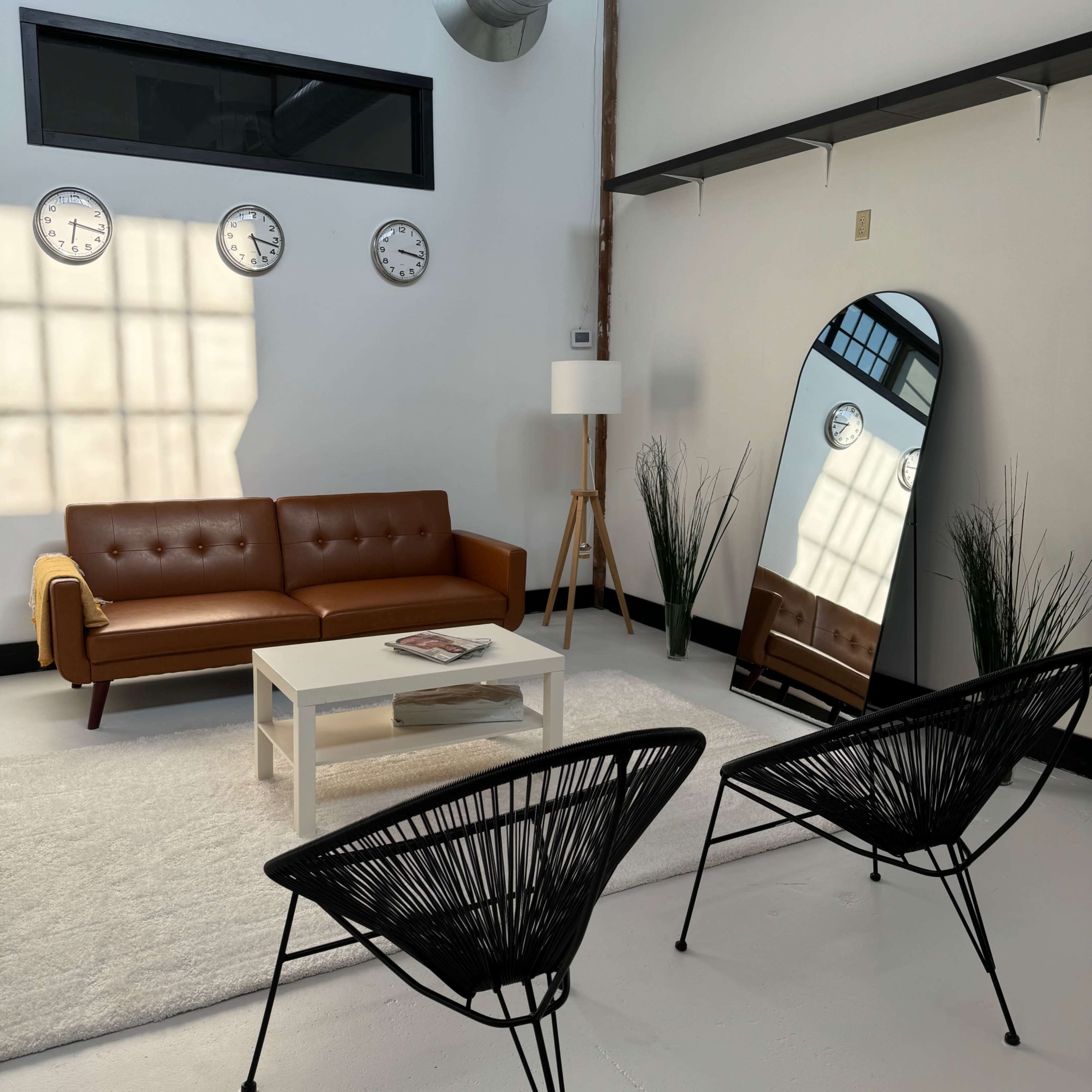 A modern living room featuring a brown leather sofa, a white coffee table, two black chairs, a large mirror, and three wall clocks, all set against a light-colored floor and walls.