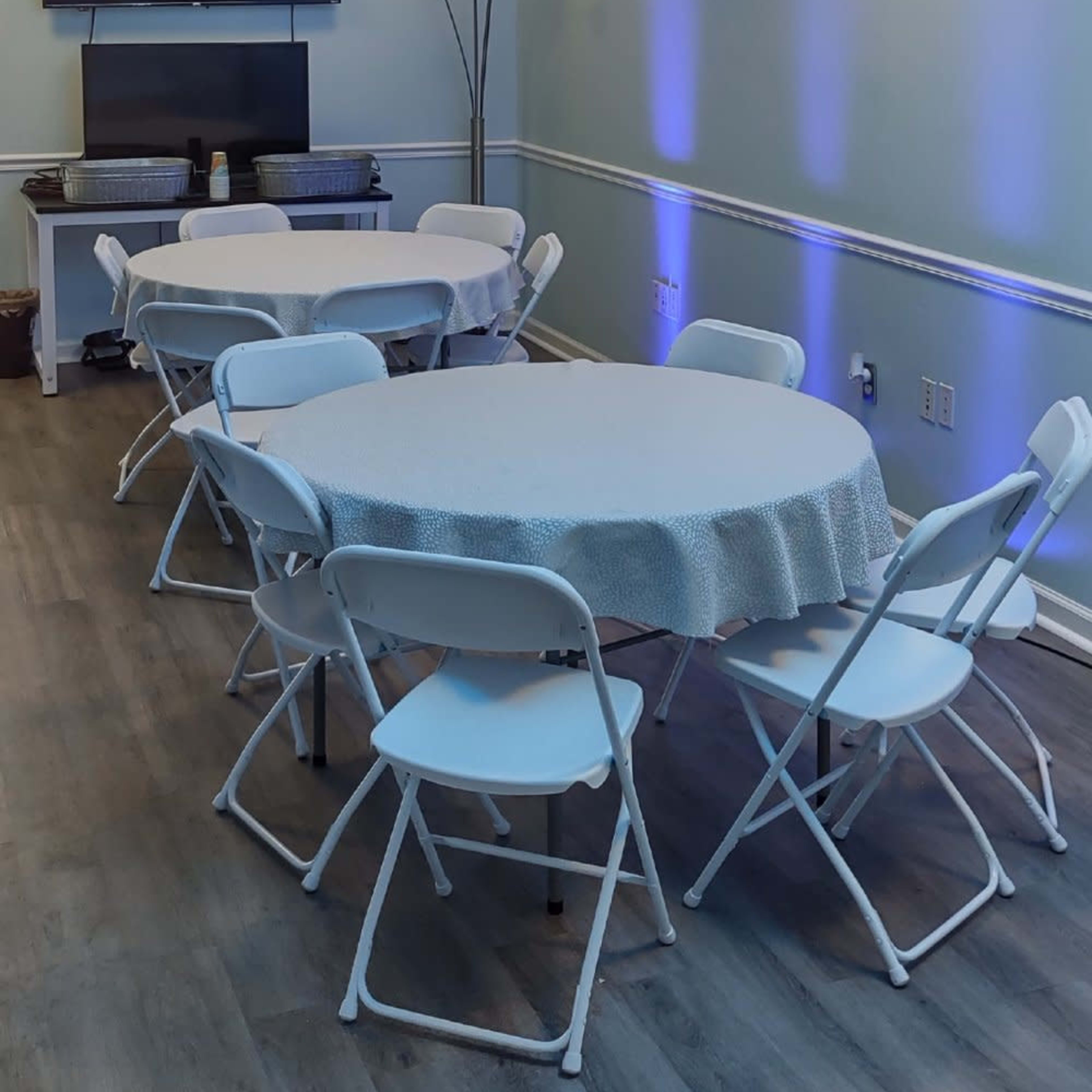 A small meeting room features several round tables surrounded by white chairs, with a television mounted on the wall and accented lighting.