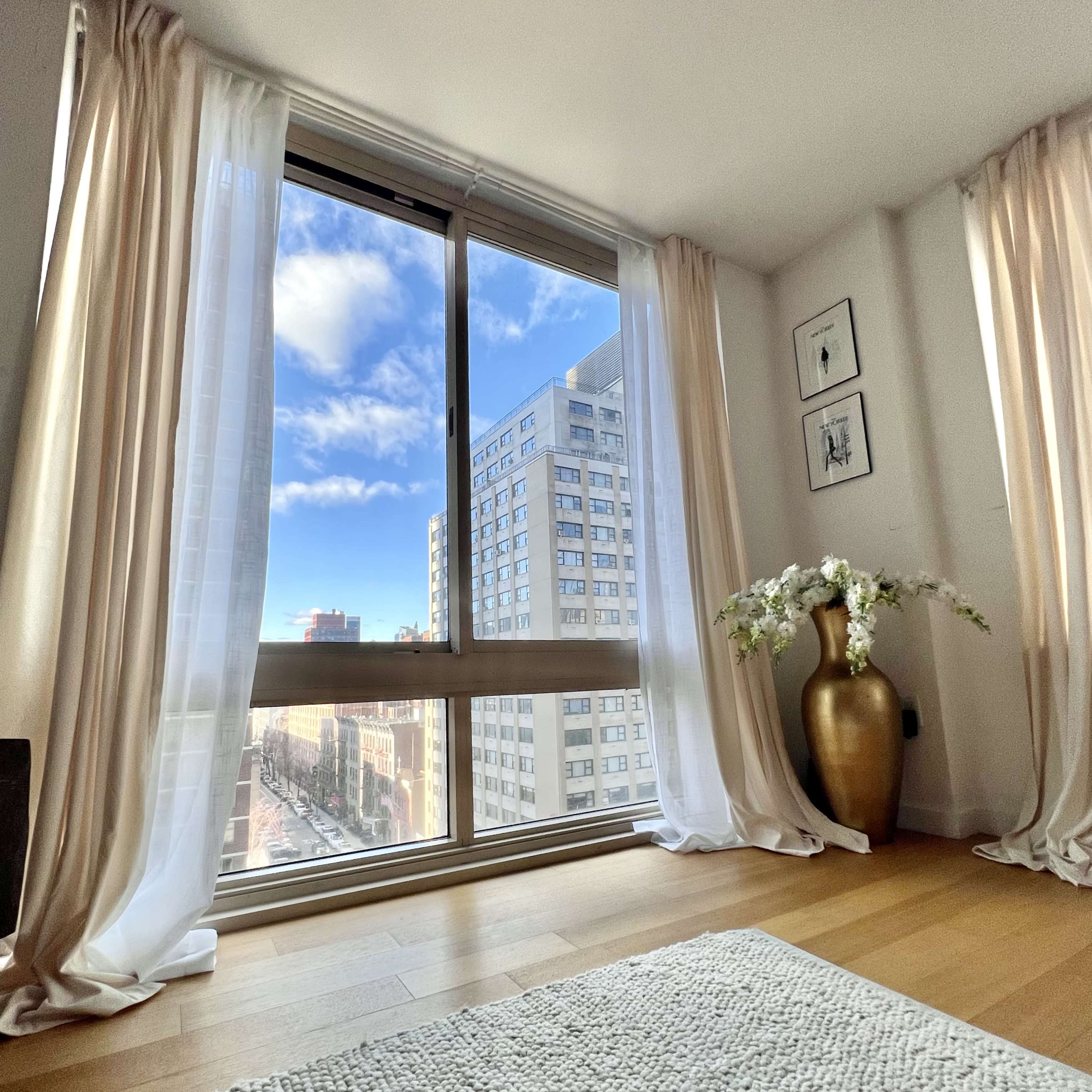 A bright room with large windows overlooks a city street, featuring sheer curtains and a decorative vase.